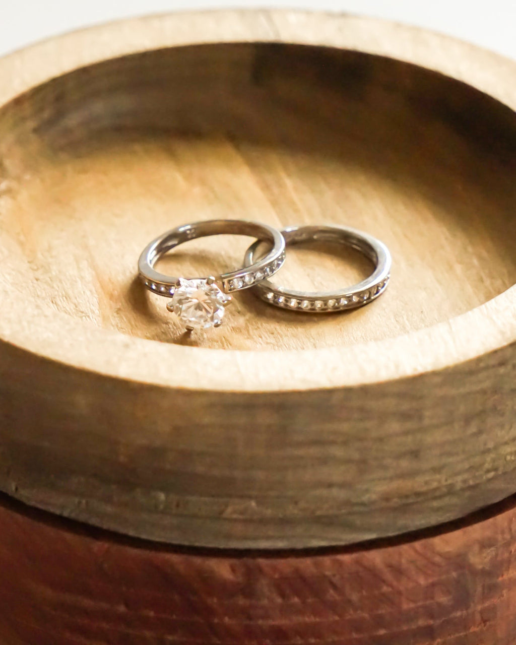 Two rings, one with a clear solitaire gemstone and the other a band of small clear stones, displayed on stacked round wooden ring dishes.