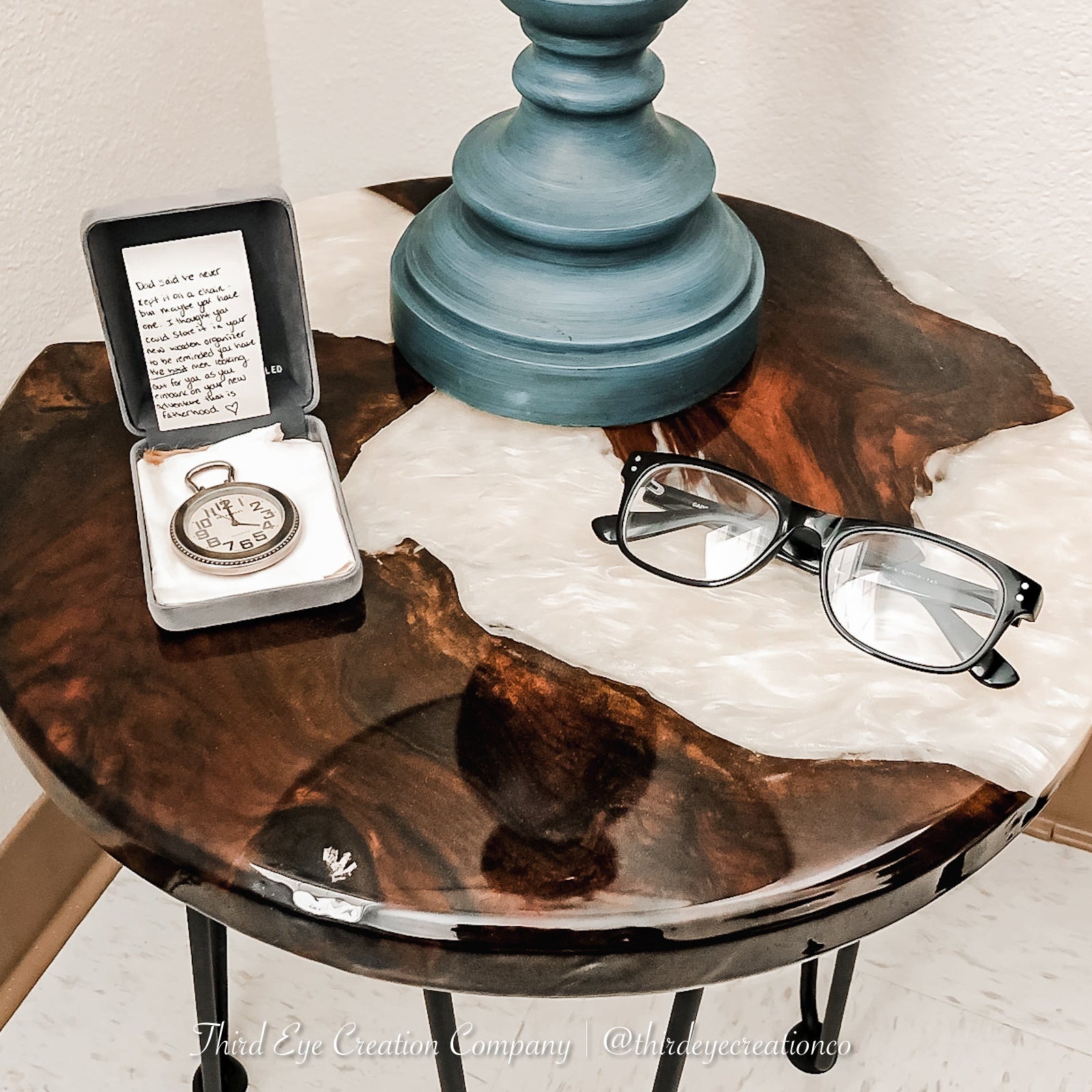 A round live-edge wood side table with a glossy dark brown wood and white resin river, styled with a teal lamp base, eyeglasses, and a pocket watch in a jewelry box.
