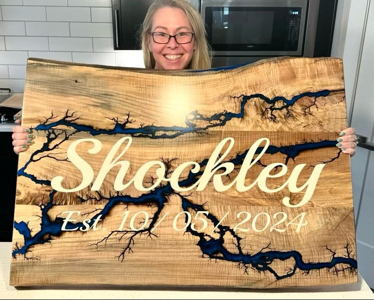 A woman in a kitchen holds up a large personalized wooden plaque featuring a live-edge wood grain design with dark blue resin river details and the name 'Shockley' in white script.