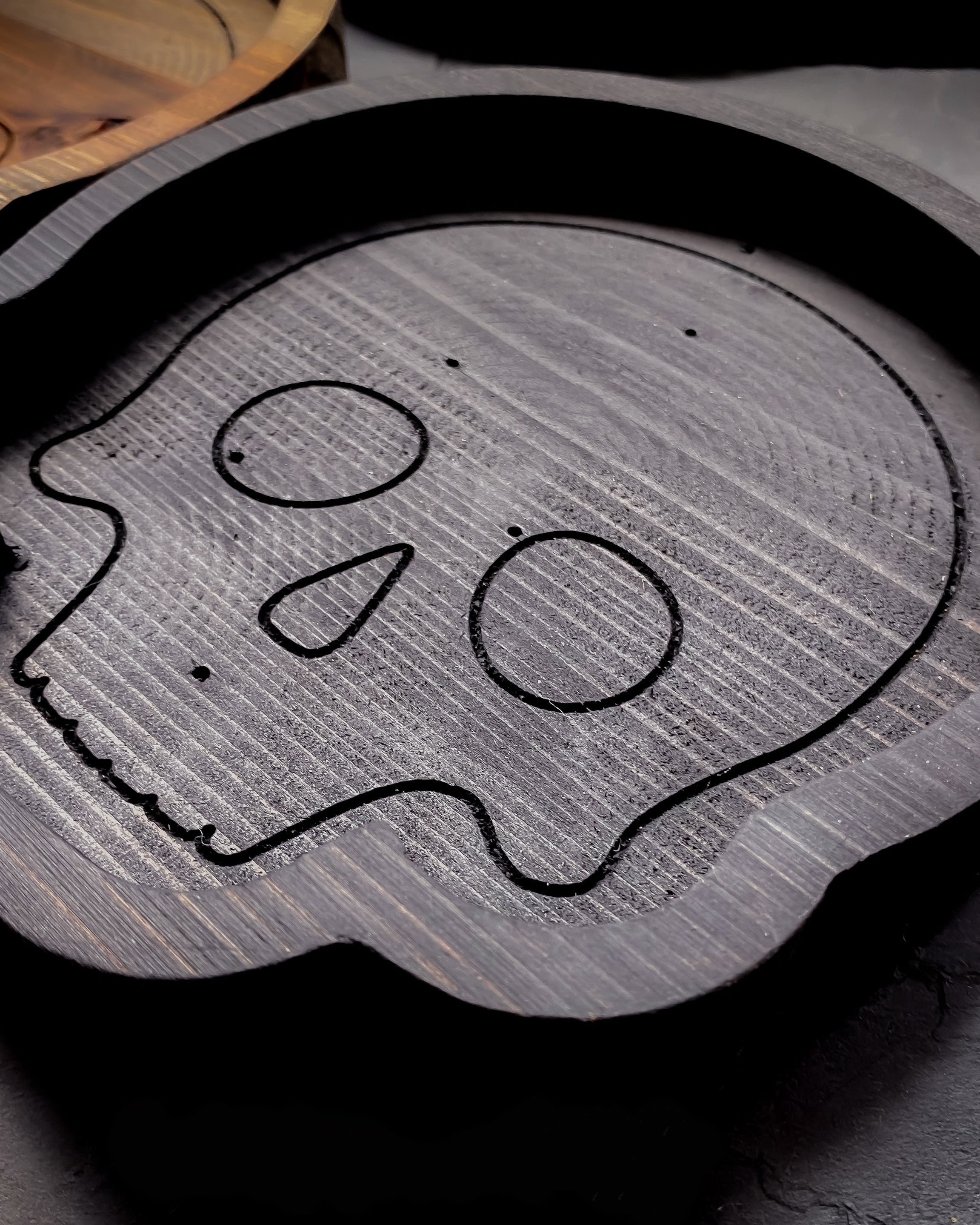 A black gray carved wooden skull-shaped tray with a hand-drawn black line art design, shown on a dark textured surface.