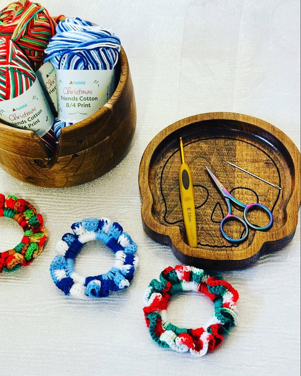 Four knitted yarn hair elastics in red, green, blue, and white striped patterns are displayed on a white textured background, with three elastics arranged outside a wooden bowl and one inside, alongside yarn skeins in a wooden container and crochet hook and scissors on a wooden skull tray.