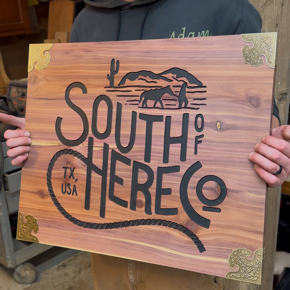A person holds a rectangular wooden sign with a warm honey-toned Cedar grain. The sign features black engraved lettering reading 'SOUTH OF TEXAS HERE CO' with a cowboy and cactus silhouette, plus brass corner brackets.