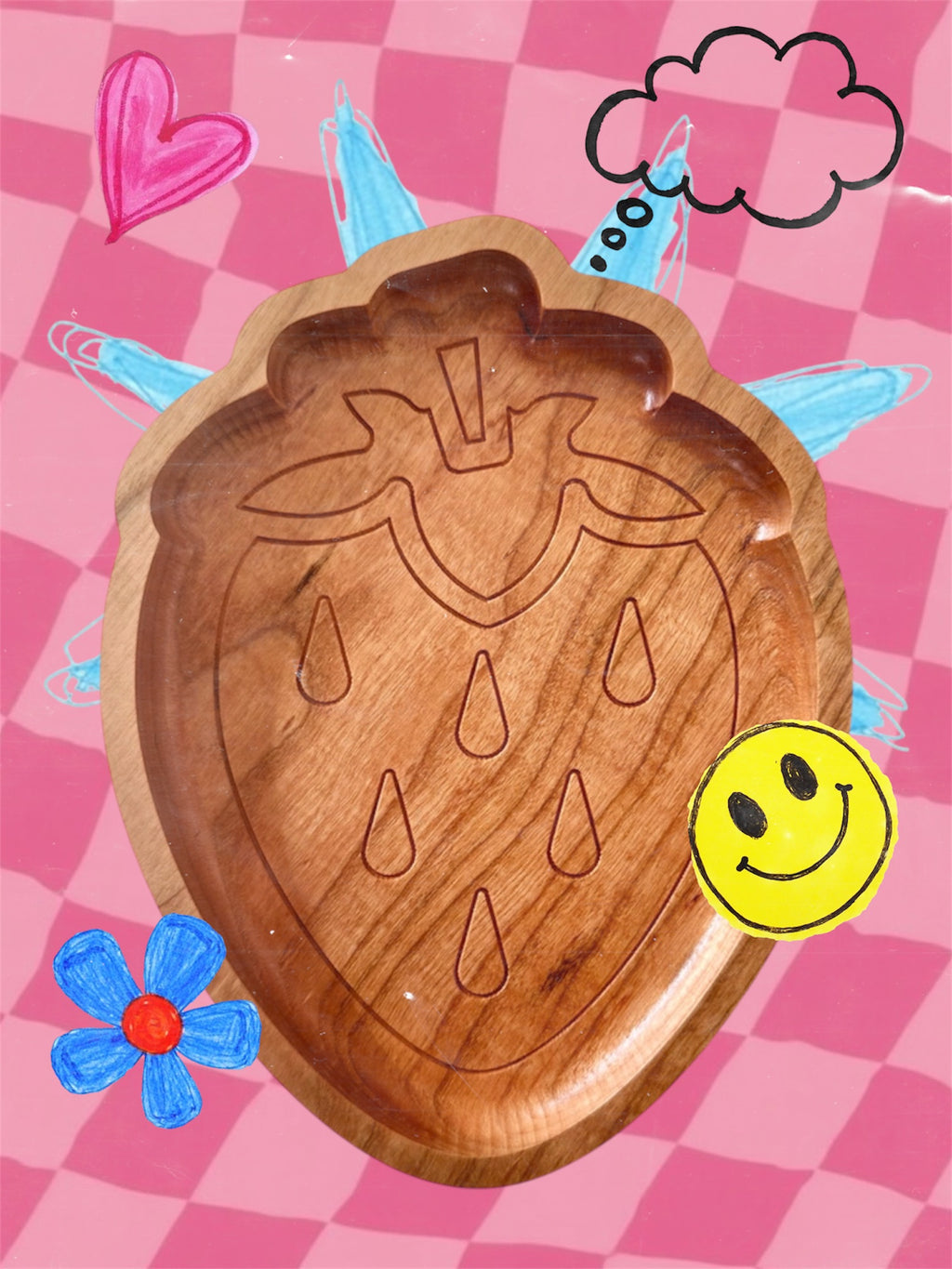 A wooden strawberry-shaped tray with carved details, displayed on a pink checkered background with hand-drawn doodles including a heart, cloud, smiley face, and flower.