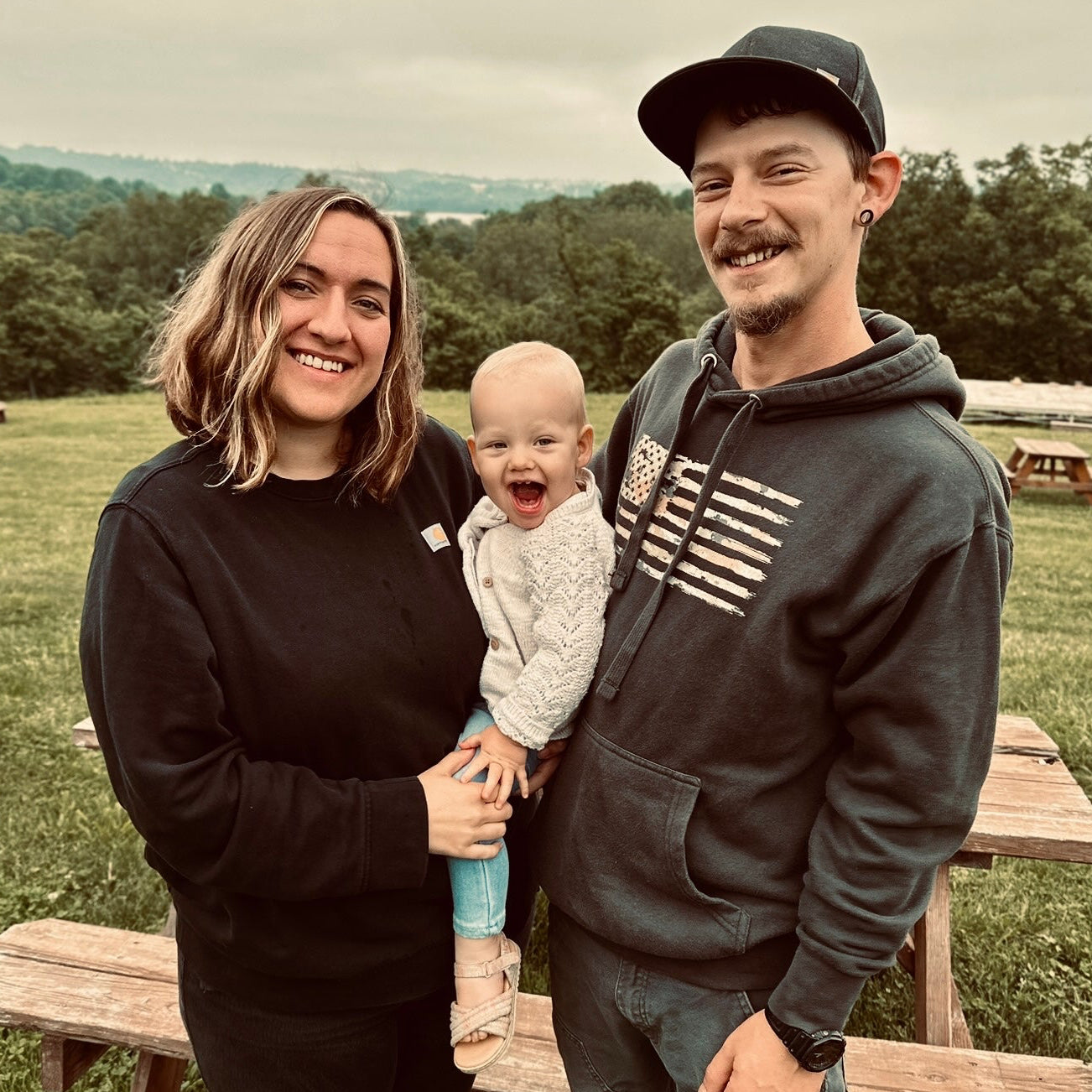 Family of three standing outdoors in a park with picnic tables and greenery.