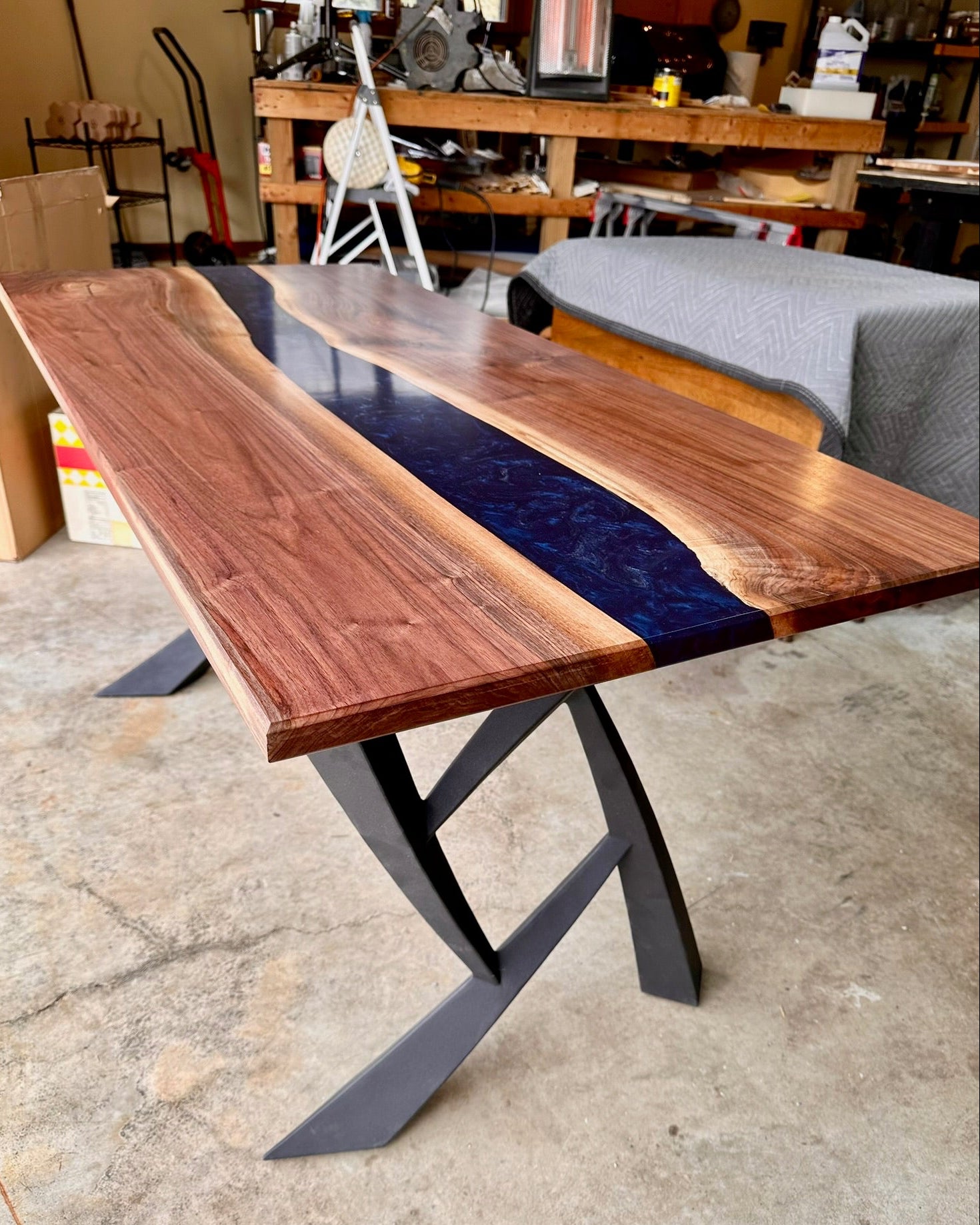 A live-edge wood table with a dark blue resin inlay and black metal X-shaped legs, photographed in a workshop environment.