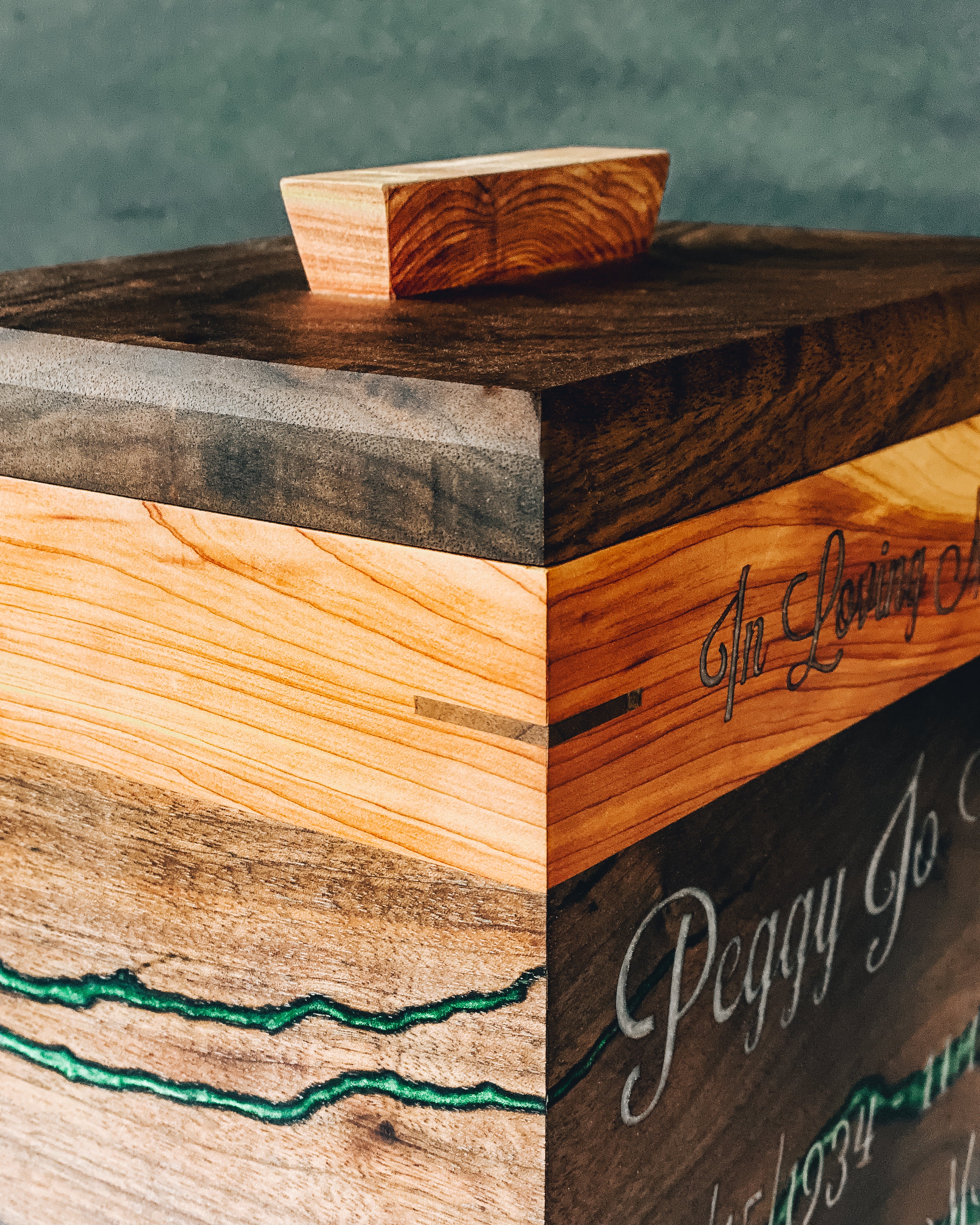 A close-up of a rustic urn box with mixed wood grain panels, a small wooden lid piece on top, and engraved script text on the side.