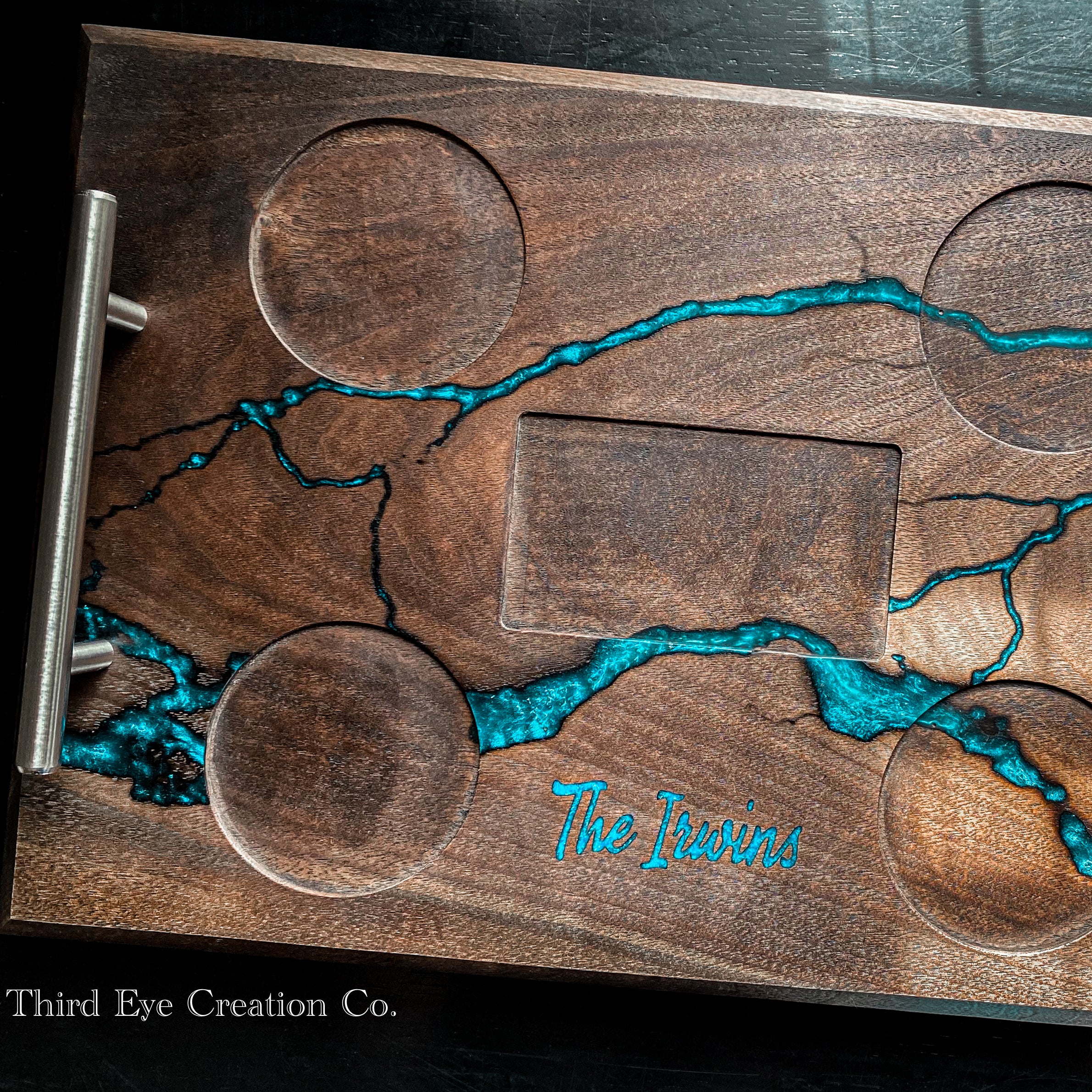 A close-up of a wooden serving tray or board with a brushed metal handle on the left. The tray has a dark brown wood grain finish with bright turquoise blue resin inlay forming a river-like pattern, circular and rectangular cutouts, and the words 'The Irwins' engraved near the bottom.