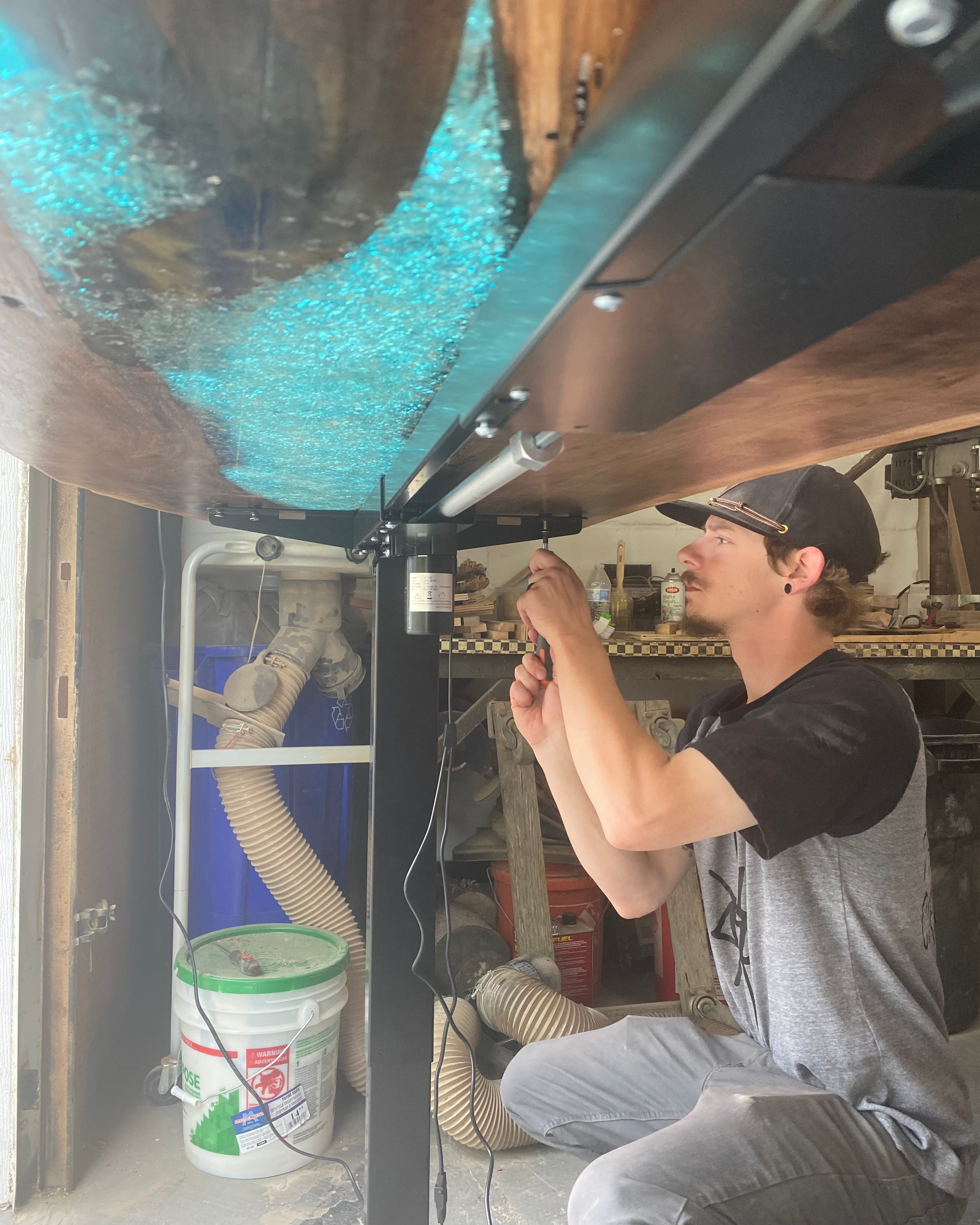 A man in a black t-shirt and grey pants is kneeling down to screw in the table legs on a large resin river table in a workshop setting.
