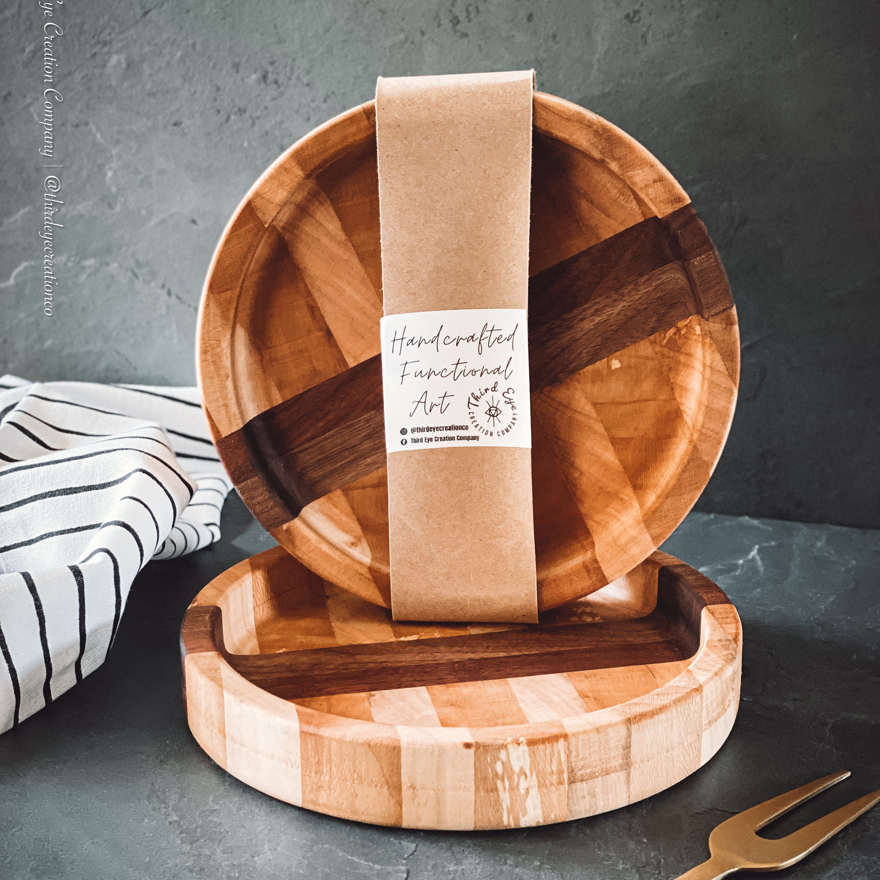Two round wooden serving bowls wrapped in a brown paper band that reads 'Handcrafted Functional Art.' The board is shown on a dark gray surface with a striped cloth and a gold-toned serving fork nearby.
