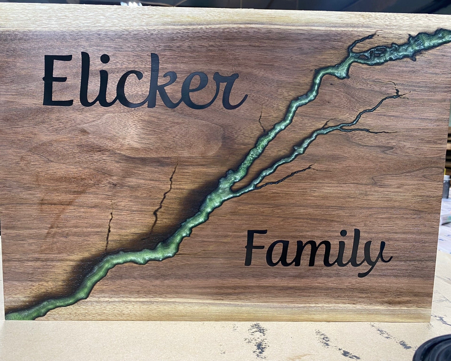 A custom wooden sign featuring a natural wood grain background with a green resin crack running diagonally across it. The words 'Elicker' and 'Family' are custom engraved in black script on either side of the crack.