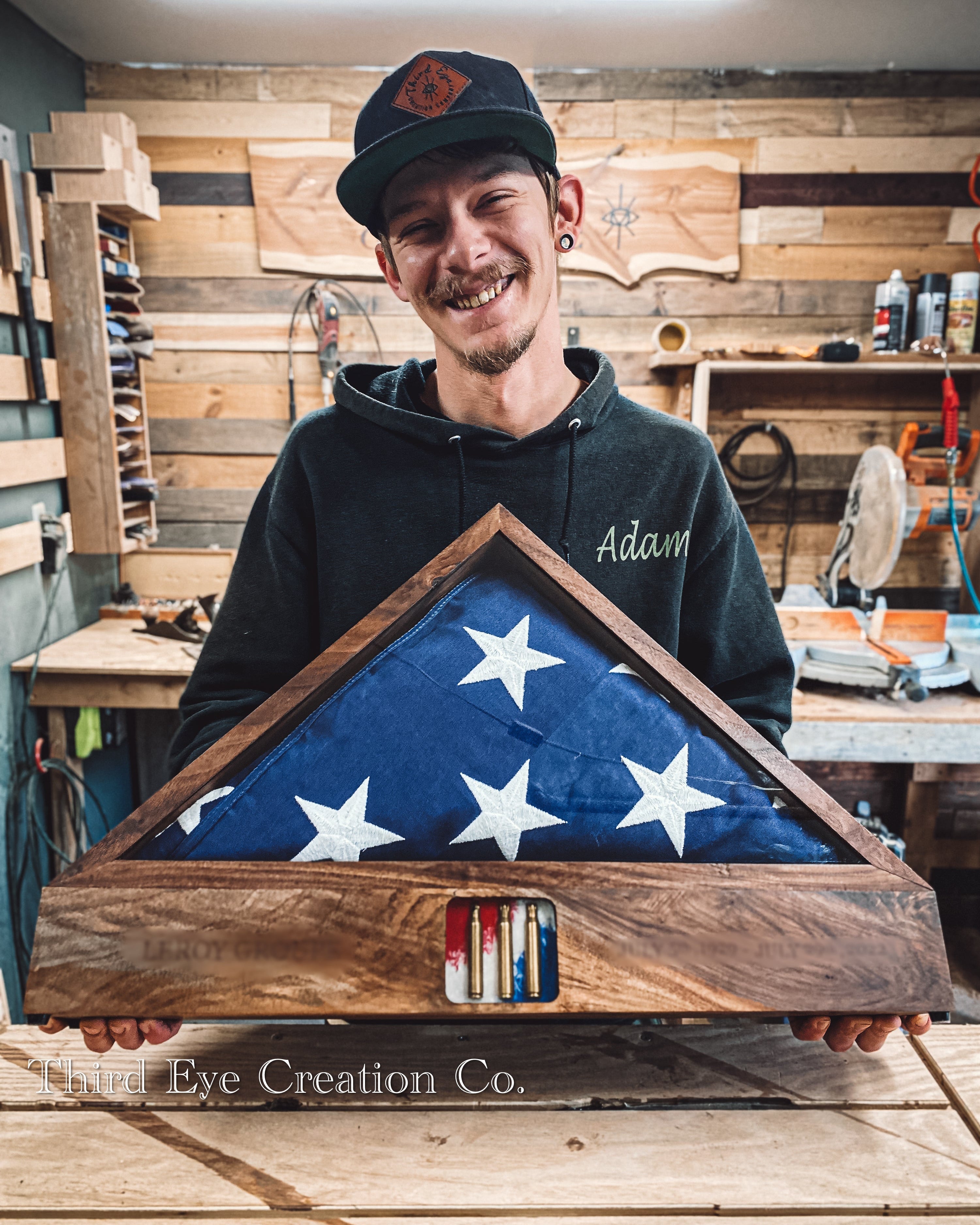 A man in a workshop holds a triangular wooden display case containing a folded American flag. The case has a dark stained wood frame and a small slot at the bottom holding bullets. 