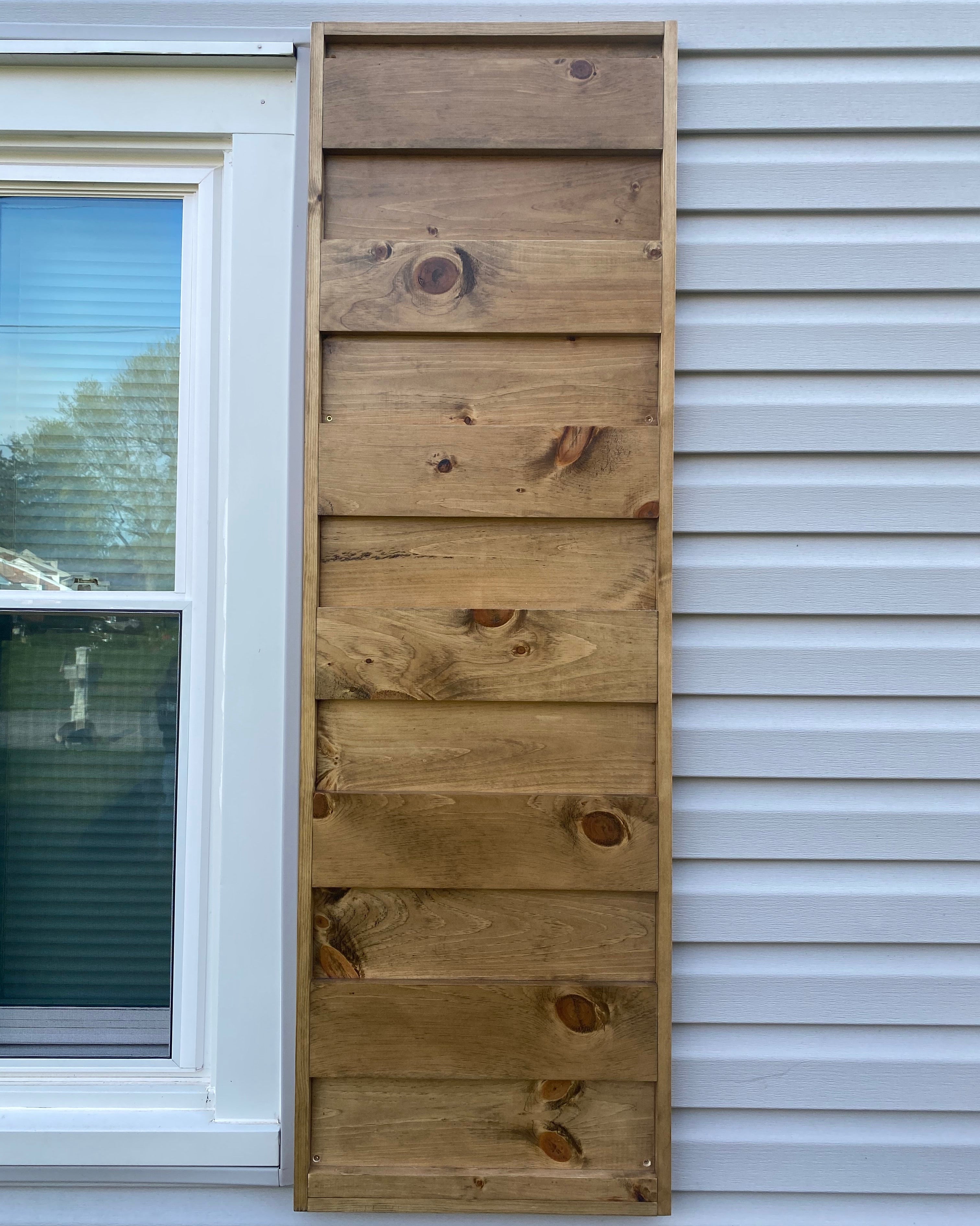 A rectangular shutter mounted on a white exterior house siding beside a window.