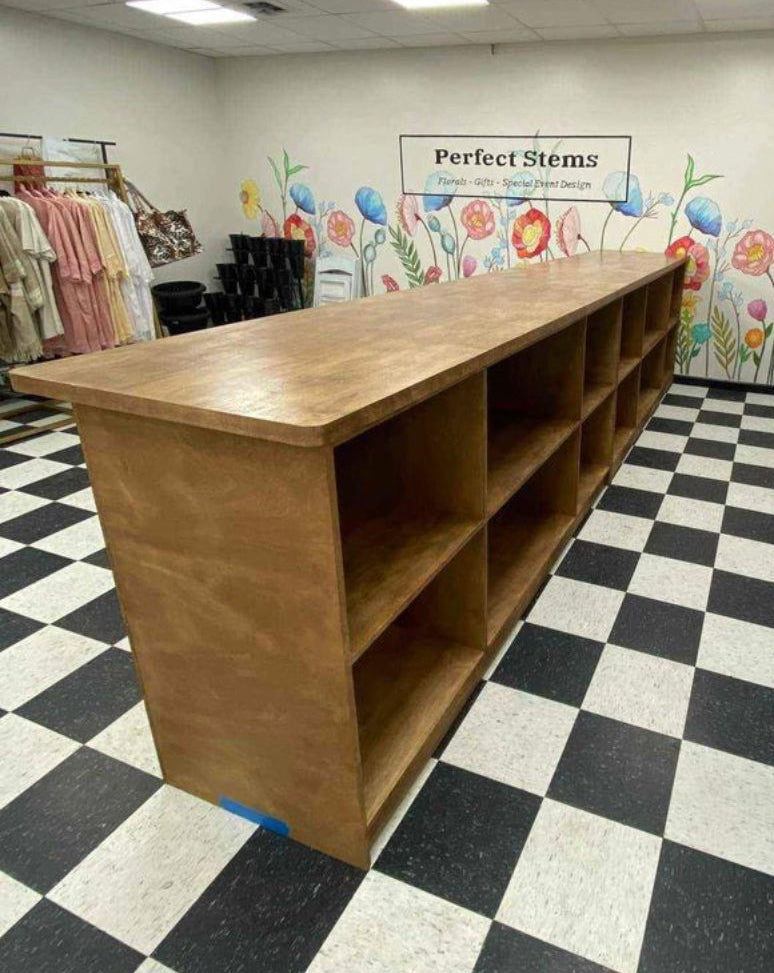 A long wooden checkout counter with multiple open cubbies, placed in a retail store on a black and white checkered floor. A colorful floral mural and a sign reading 'Perfect Stems' are visible on the back wall.