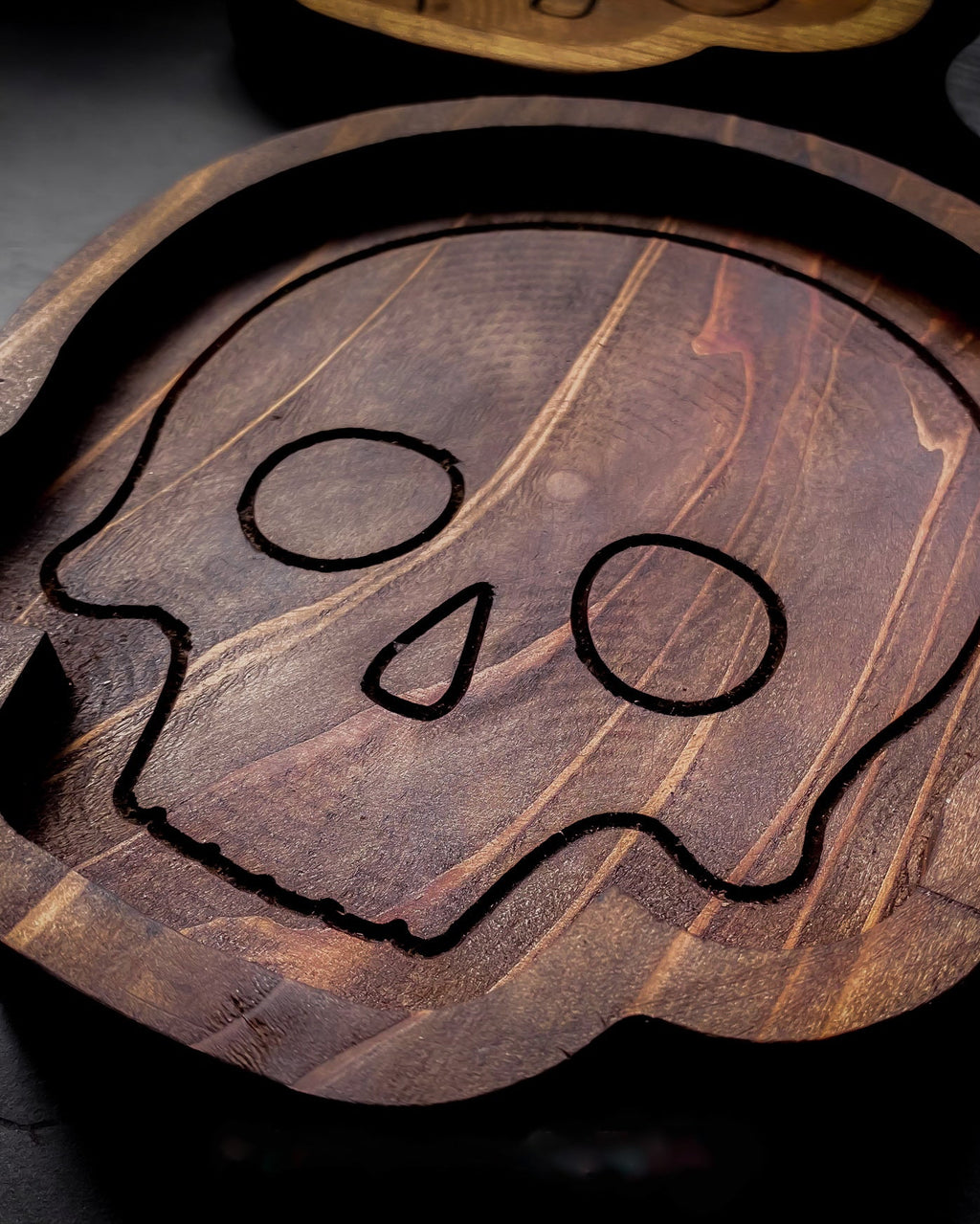 A dark brown wooden skull-shaped tray with engraved black line art of a skull, shown on a dark textured surface.