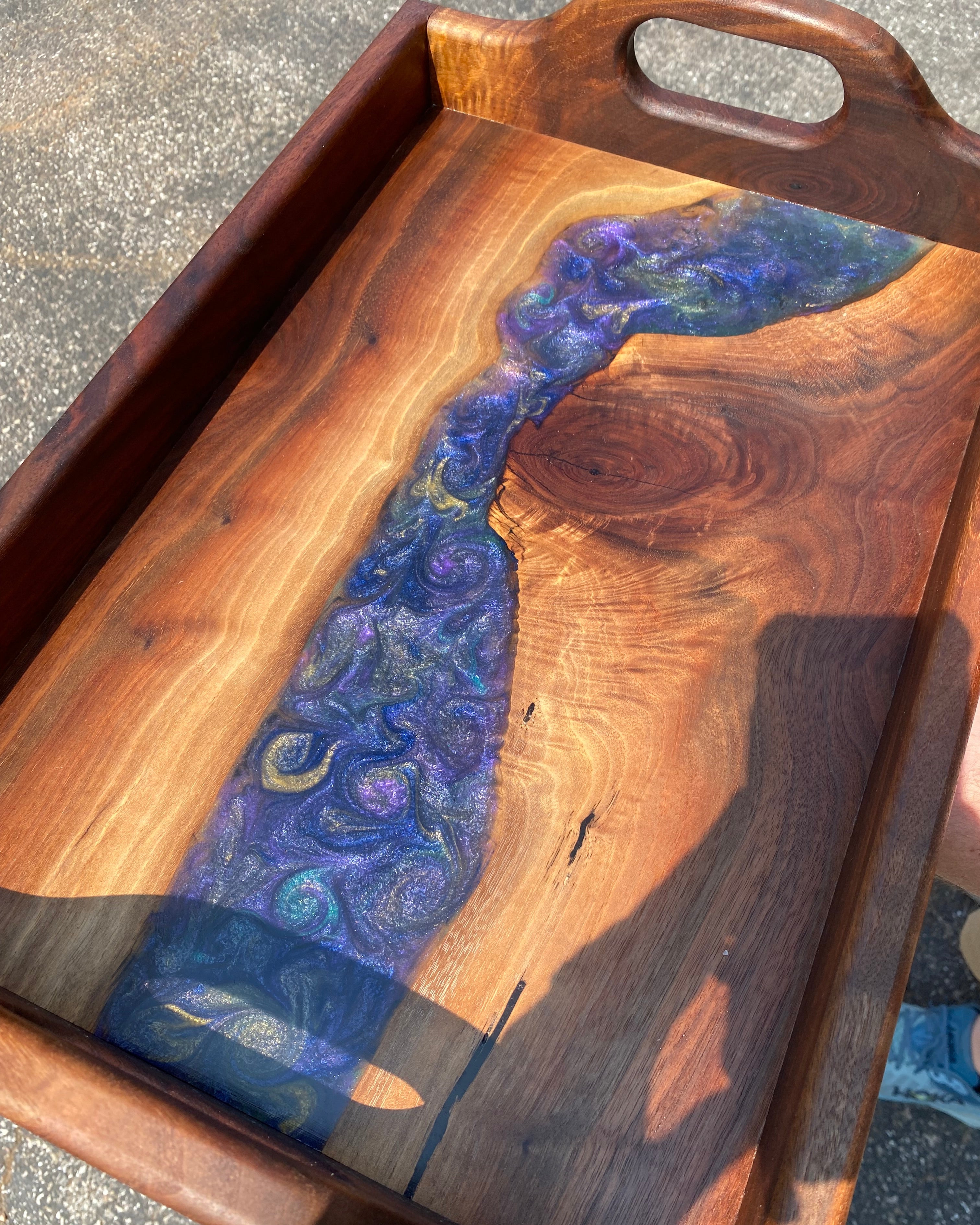 A rectangular walnut wooden serving tray with a live edge in the center handles cut on the sides, featuring a swirling purple and blue resin inlay over a natural wood grain surface.