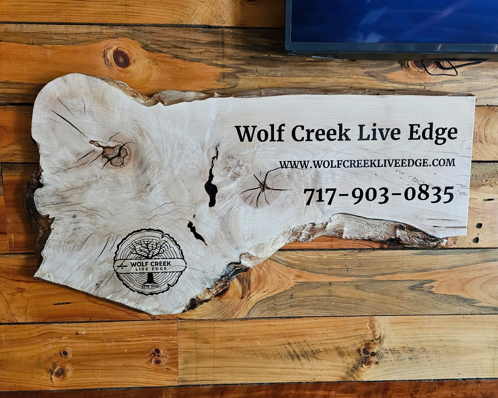 A rustic live-edge wood sign mounted on a wood plank wall, featuring the text 'Wolf Creek Live Edge' with website and phone number printed on a white section, plus a circular Wolf Creek logo stamp.