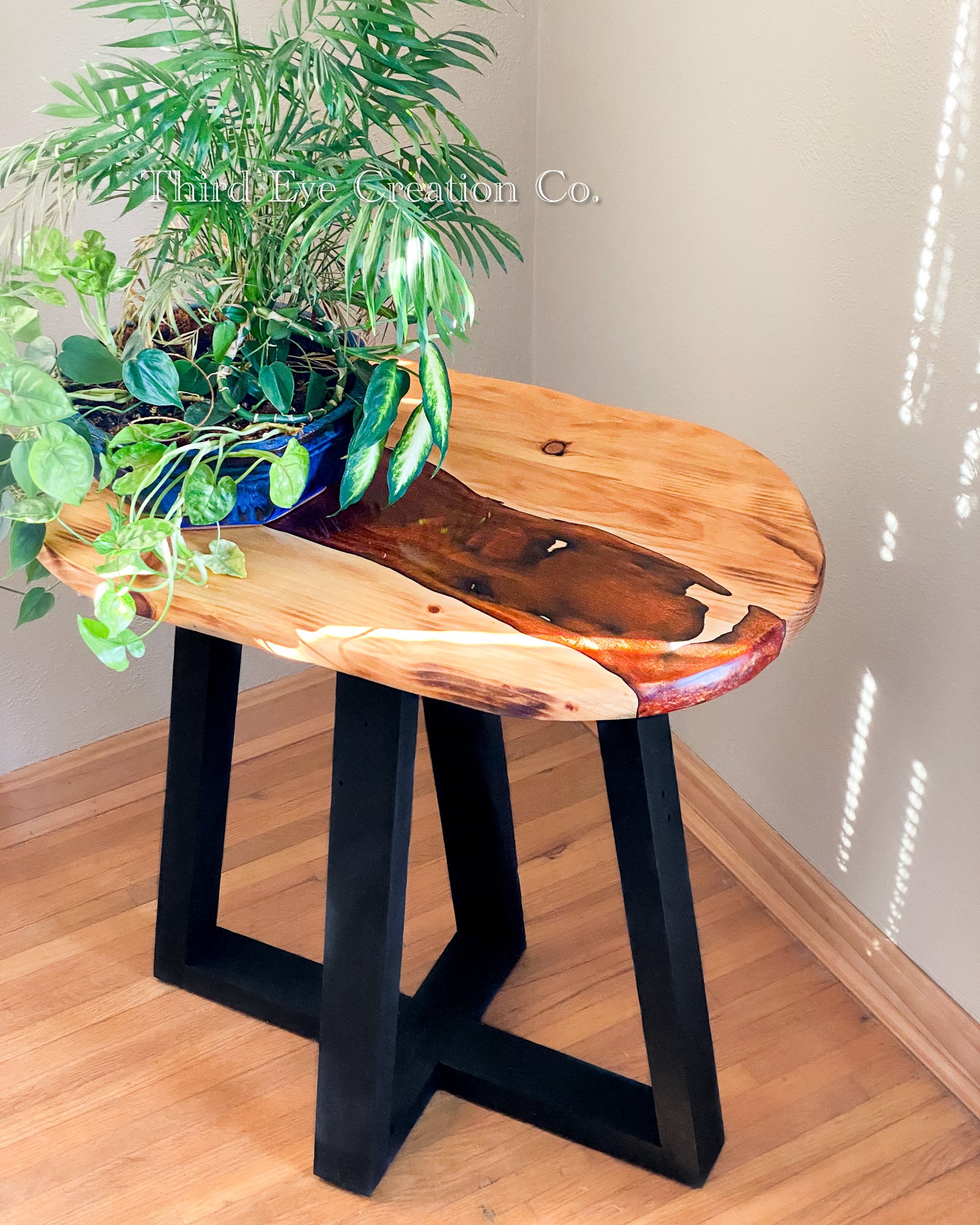 A rustic live-edge wooden table with a rustic orange/red river and black wood base, styled with a potted green plant in a room with light wood floors and beige walls.