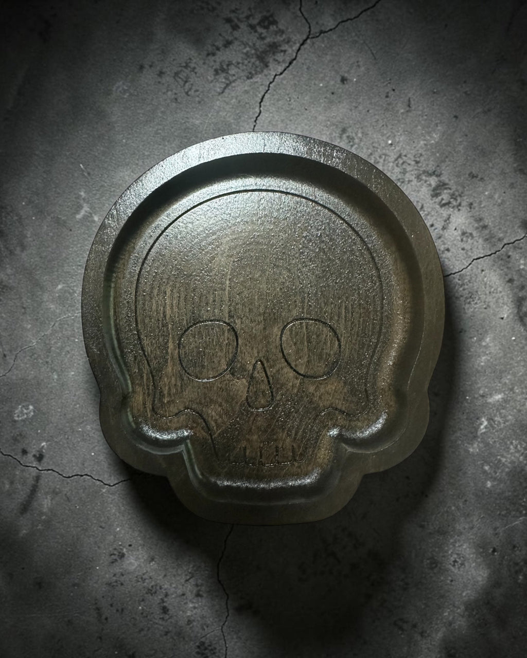 A skull-shaped wood tray with a textured, weathered finish, set against a dark, cracked concrete background.