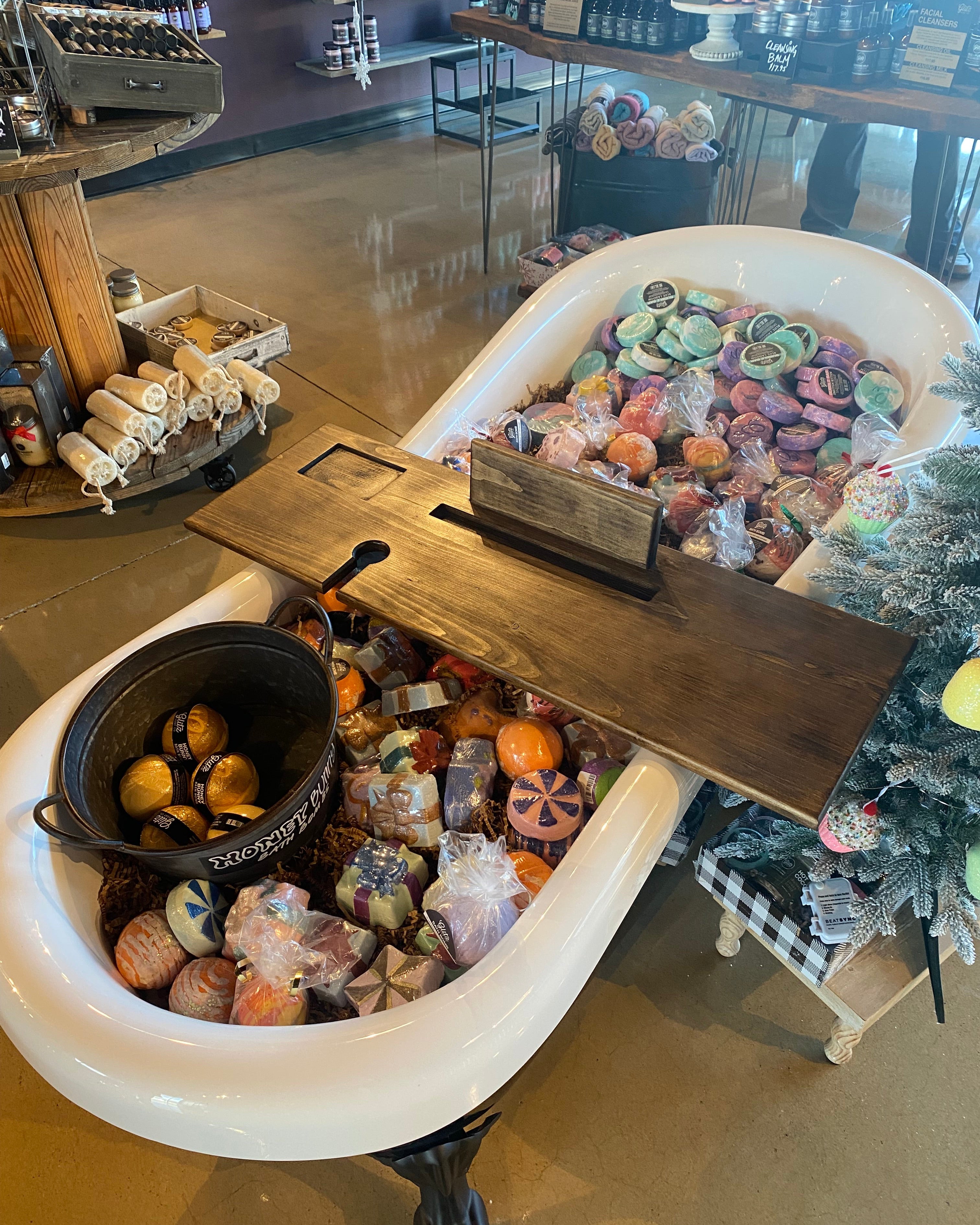 A large white bathtub display filled with assorted decorative bath products, including a brown bath caddy, colorful bath salts, wrapped soaps, and a black bucket of bath bombs, photographed in a retail store setting.