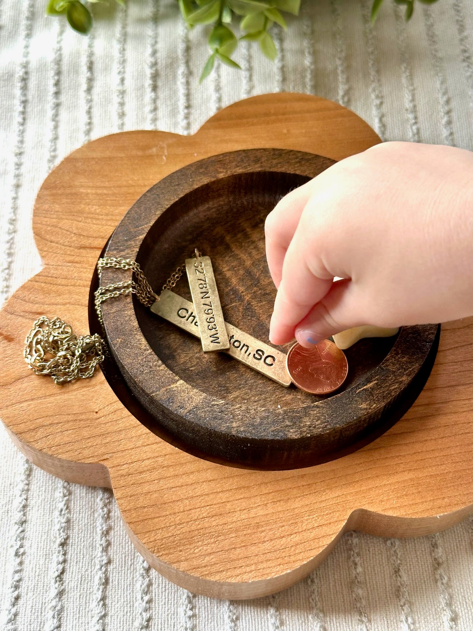 A hand placing a penny into a flower-shaped wooden money tray with engraved gold-tone nameplates, alongside a gold-tone chain necklace, on a textured white fabric background.