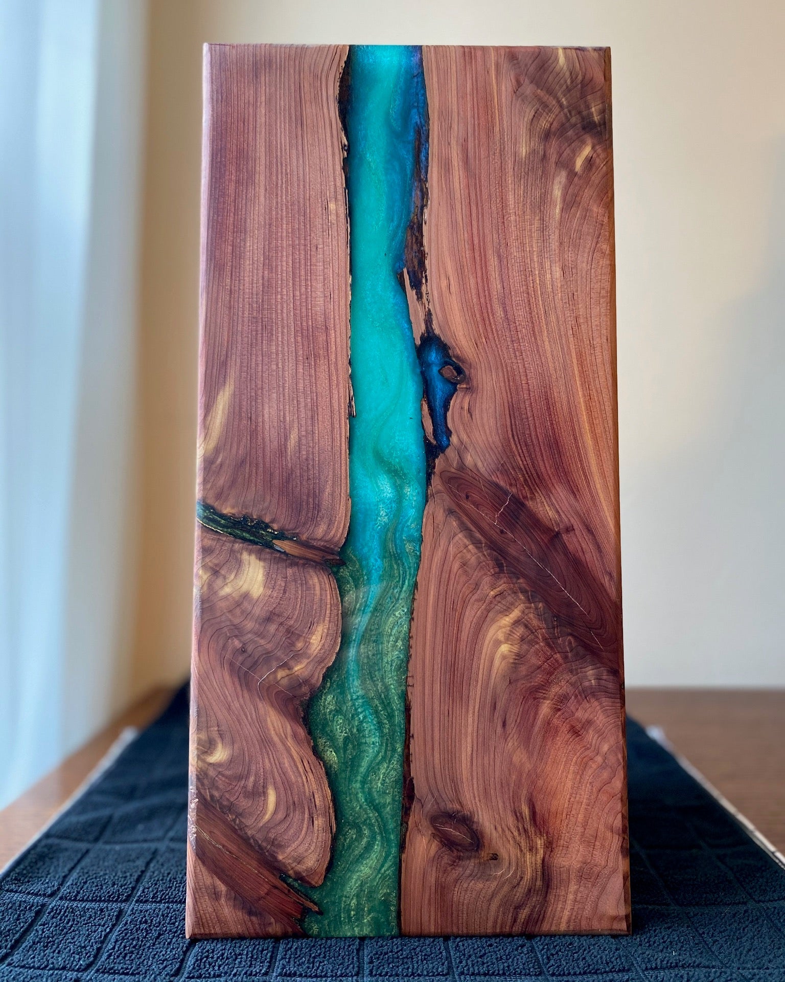 A rectangular cedar slab art piece with a vibrant turquoise and deep blue resin river running through a natural wood grain pattern, standing upright on a dark textured surface.