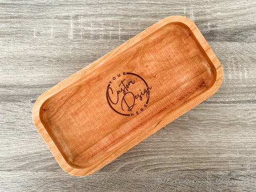 A rectangular wooden tray with rounded corners and a laser-engraved logo in the center, displayed on a gray wood-textured background.