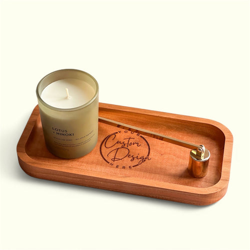A wooden tray holding a lit candle in a clear glass jar and a brass scented candle holder, photographed on a clean off-white background. The candle label reads LOTUS + MINKI, and the tray is engraved with a logo that says Custom Design Here.