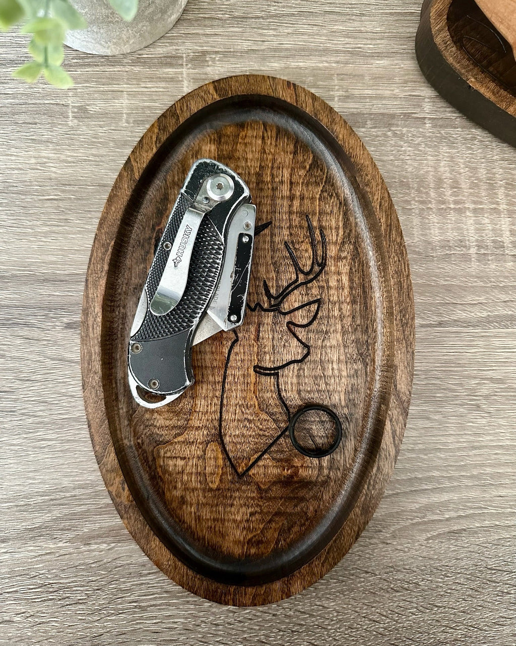 An oval wooden tray with a carved deer head design, holding a folding pocket knife and a black ring on a gray wood-grain tabletop.
