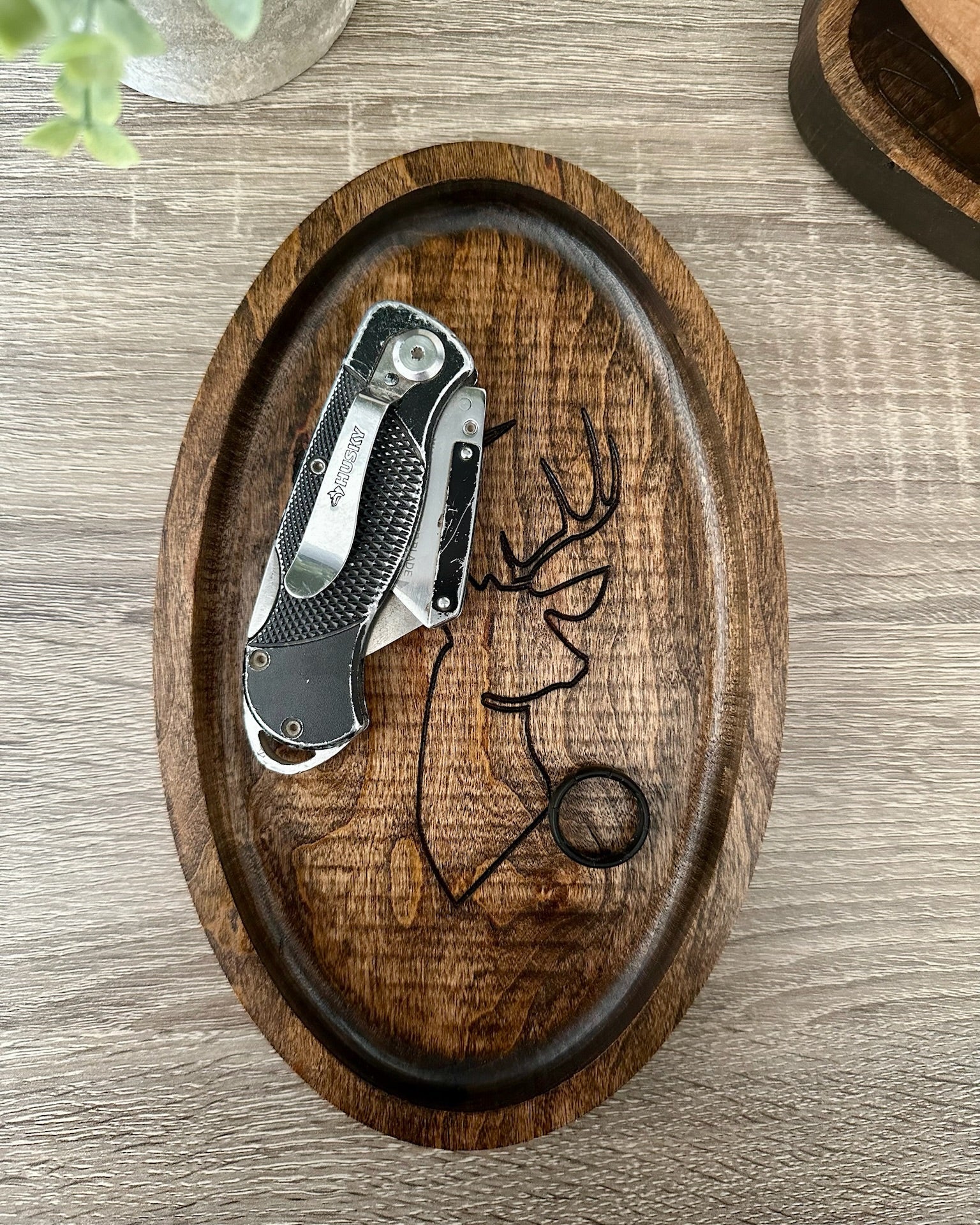 An oval wooden tray with a carved deer head design, holding a folding pocket knife and a black ring on a gray wood-grain tabletop.