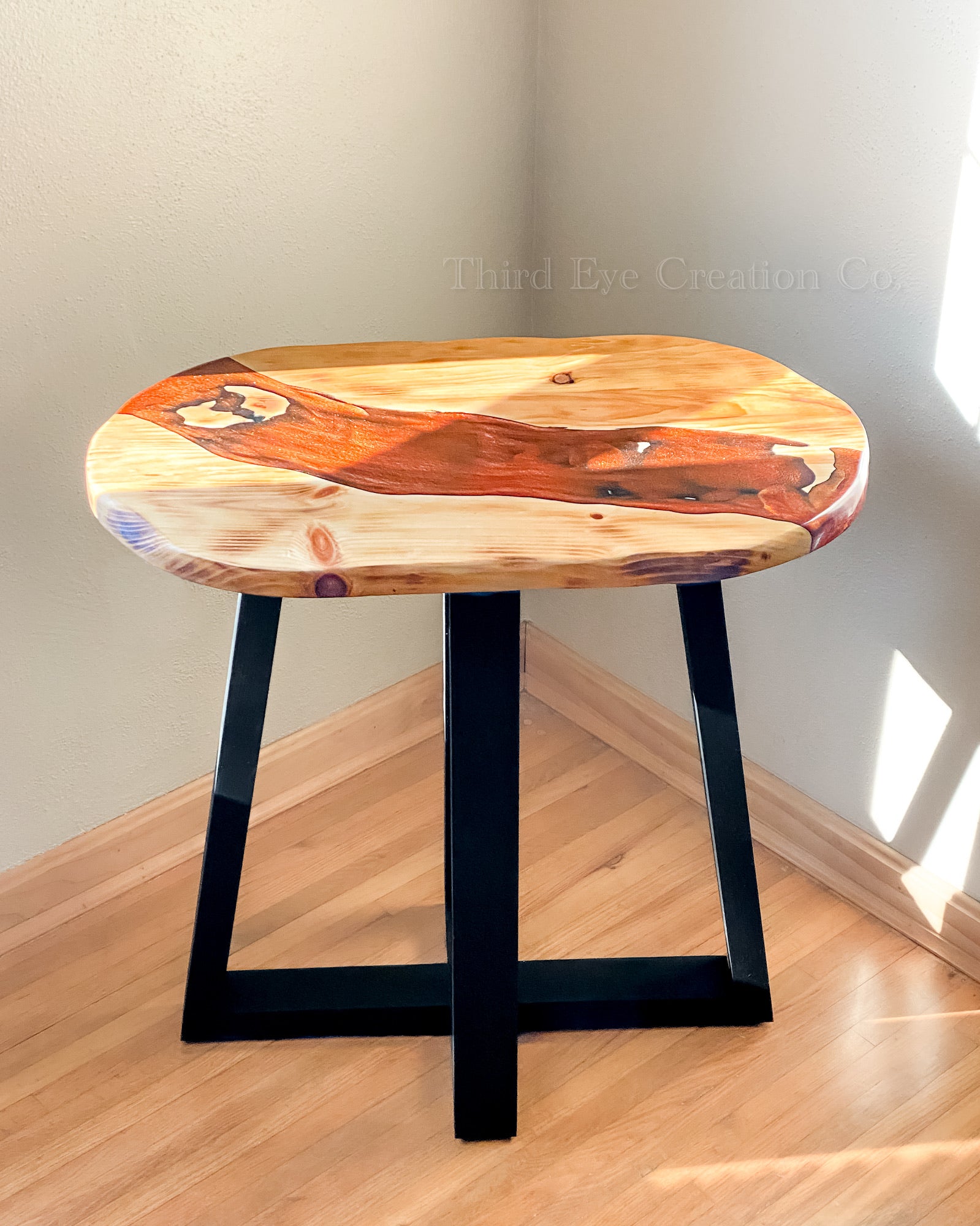 A rustic live-edge wood stool with a polished natural wood top and black metal legs, set in a room corner with light-colored walls and a wood floor.