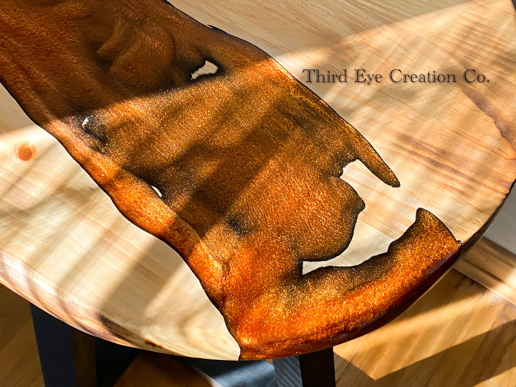 A close-up of a polished wooden tabletop with a rich amber-brown resin inlay, resting on a light natural wood surface. The text 'Third Eye Creation Co.