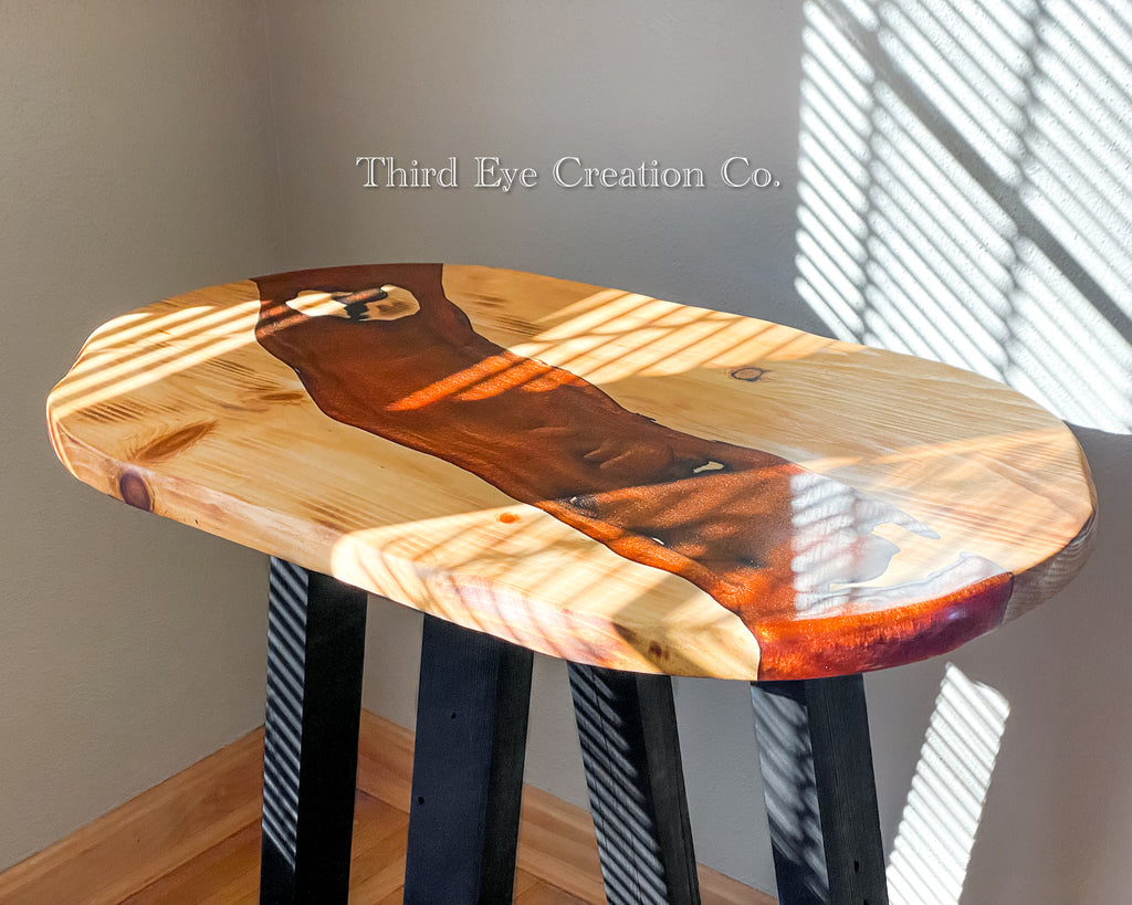 An oval live-edge wooden table with a glossy rust colored resin river and black legs, photographed in a room with sunlight casting window-blind shadows on the tabletop and wall. The watermark 'Third Eye Creation Co.