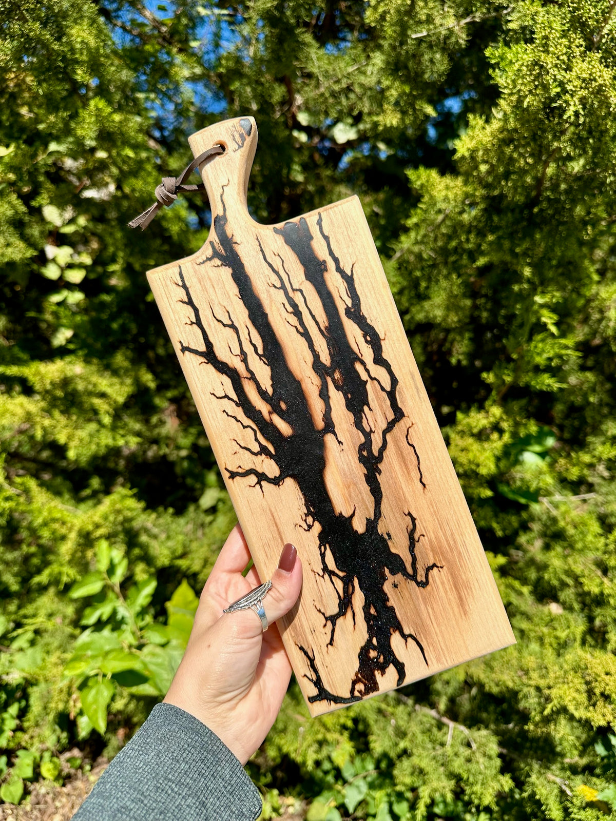 A hand holds a rectangular wooden cutting board outdoors against a background of green trees. The board has a natural light wood grain with a prominent black burned or etched branch-like pattern running vertically and branching across the surface.