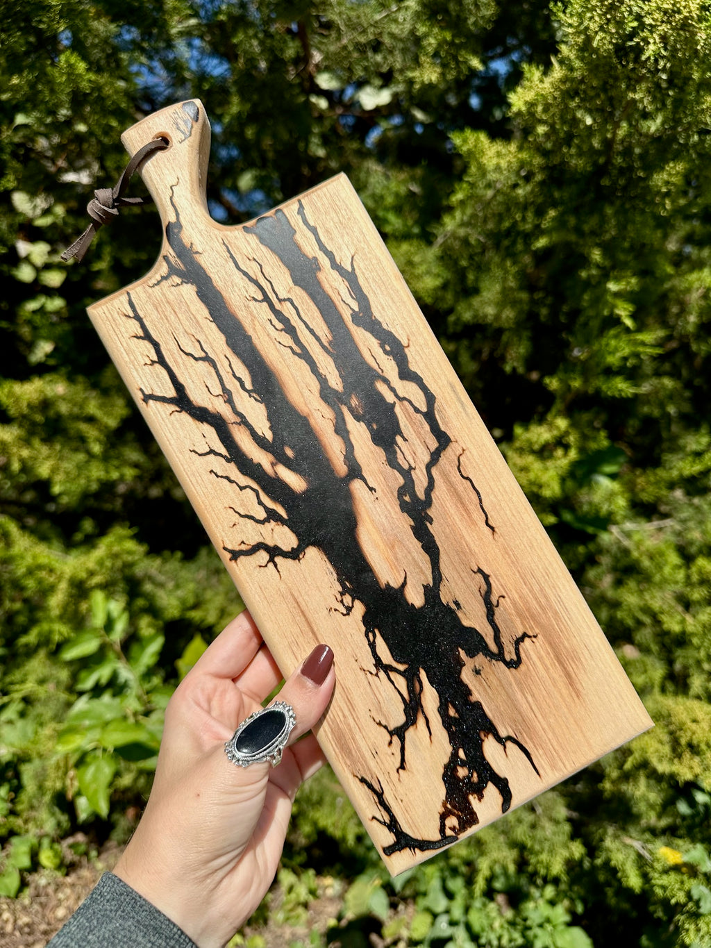 A hand holds a rectangular wooden cutting board outdoors, featuring a natural wood grain with a prominent black resin river running through the center and a dark handle.