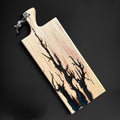 A rectangular wooden serving board with a natural wood grain finish and a dark, branch-like resin inlay running diagonally across the surface. The board features a handle with a leather hanging loop and a small decorative knot, shown on a dark background.
