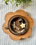 A flower-shaped wooden tray holding a smaller round wooden dish with a gold ring, a cream star-shaped object, and two engraved wooden tags, styled on a textured white fabric with green leaves at the top.