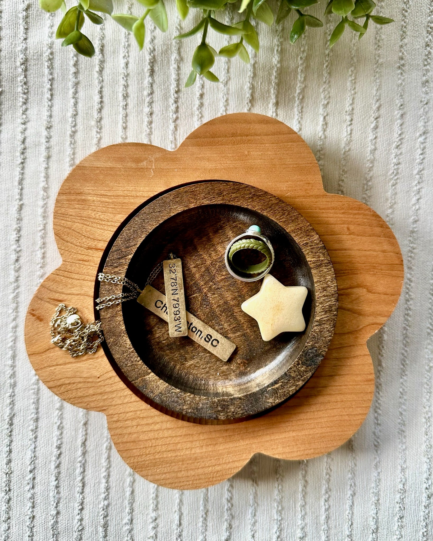 A flower-shaped wooden tray holding a smaller round wooden dish with a gold ring, a cream star-shaped object, and two engraved wooden tags, styled on a textured white fabric with green leaves at the top.