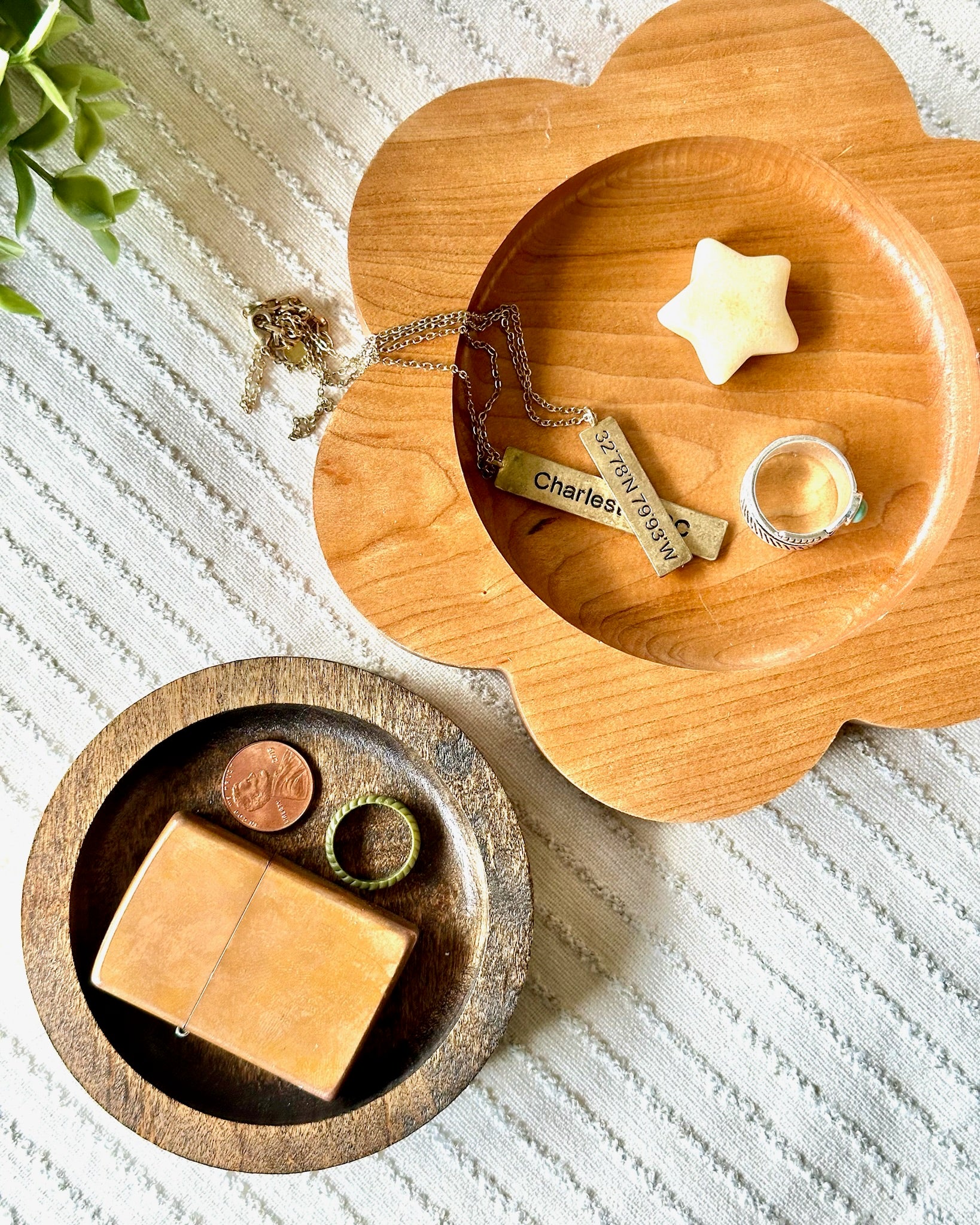 Two wooden trays, one flower-shaped and one round, styled with various small accessories including rings, a star-shaped object, a coin, and a rectangular tan item on a textured white blanket.