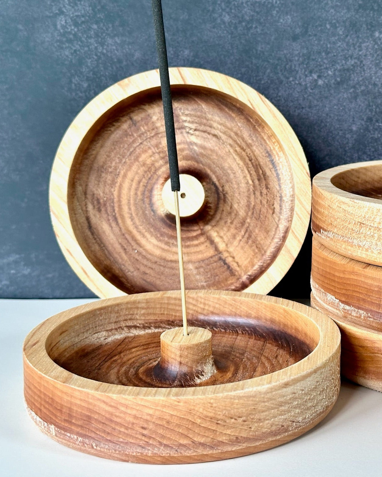 Three round wooden incense holders with visible wood grain, arranged on a light surface against a dark textured backdrop. One holder is open with a vertical incense stick inserted, and two more holders are stacked on the right.