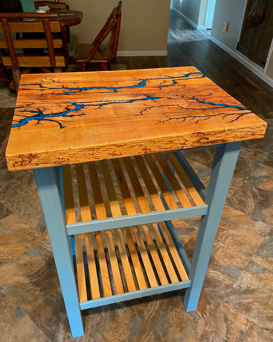 A rustic wooden end table with a live edge, featuring a warm honey-orange wood top with a blue resin fractal burned pattern, supported by a light blue frame with a slatted lower shelf, set on a stone tile floor.