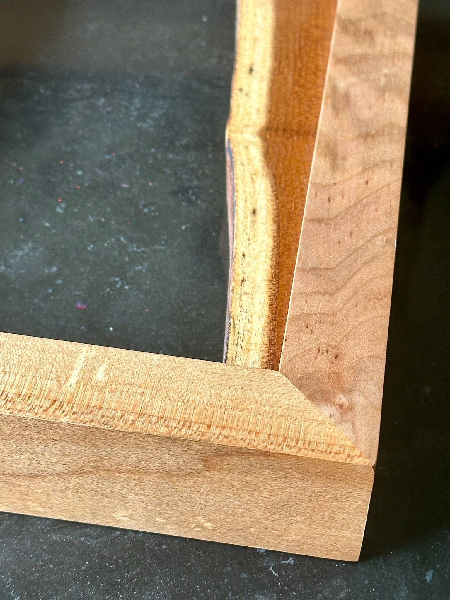 Close-up of a wooden picture frame corner showing the natural grain of light brown wood on a dark surface.