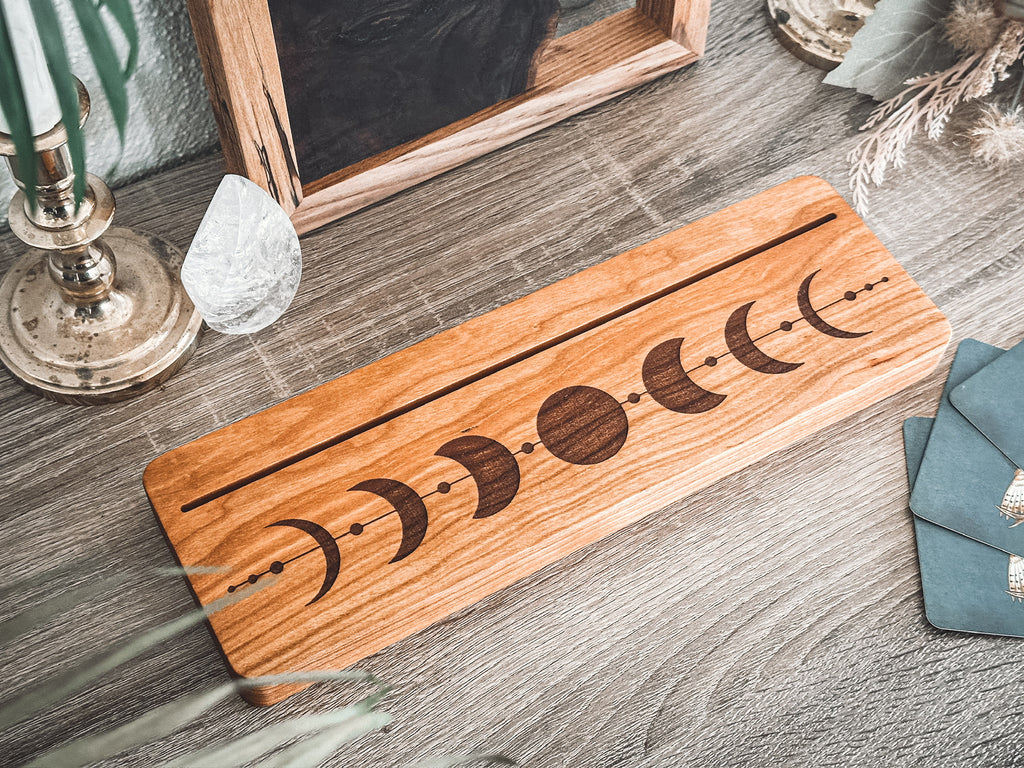A rectangular wood piece with a moon phase design engraved on it, displayed on a gray wood-textured surface with decorative props including a crystal, candlestick, and teal fabric.