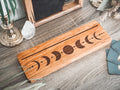 A rectangular wood piece with a moon phase design engraved on it, displayed on a gray wood-textured surface with decorative props including a crystal, candlestick, and teal fabric.