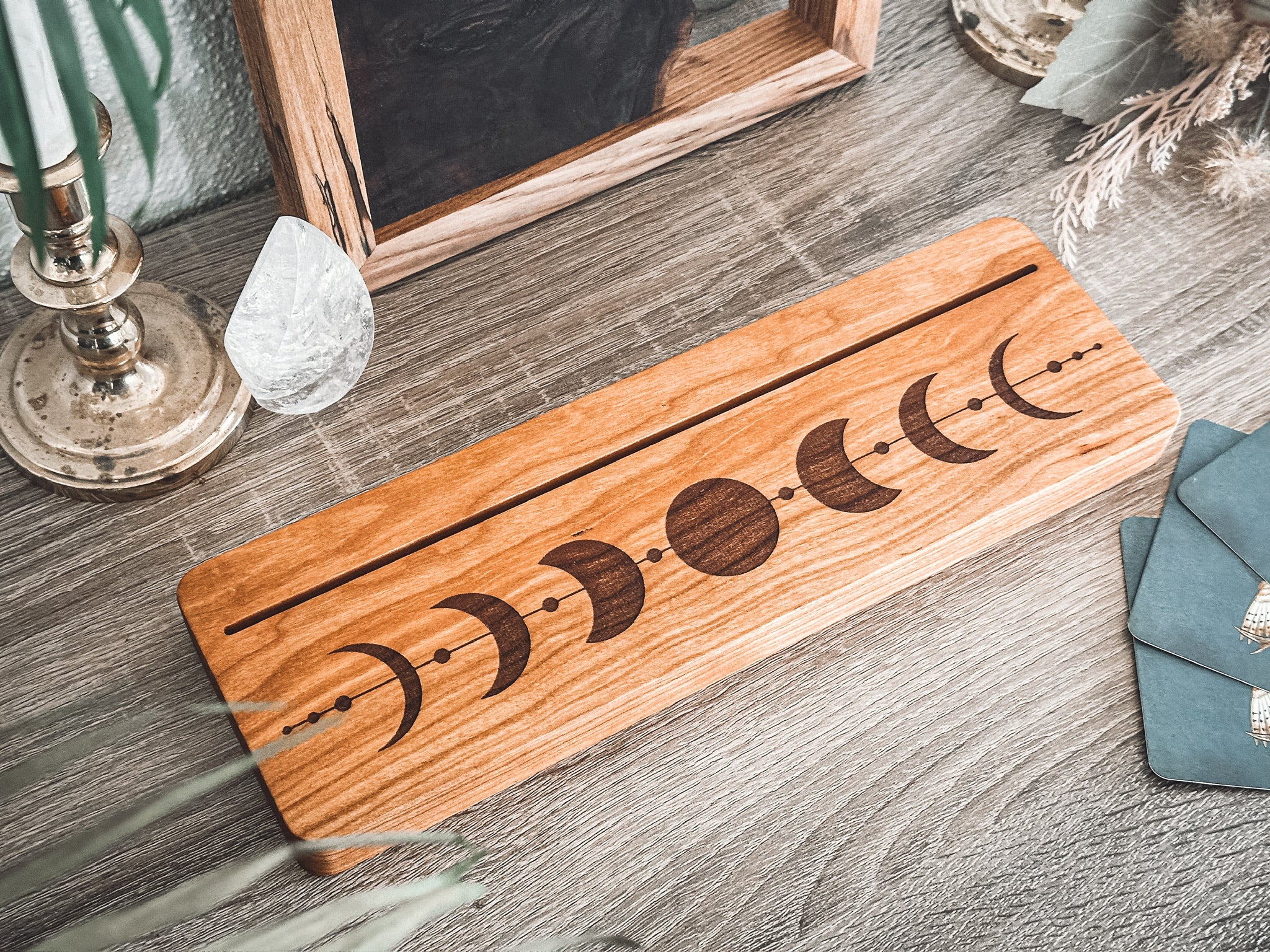 A rectangular wood piece with a moon phase design engraved on it, displayed on a gray wood-textured surface with decorative props including a crystal, candlestick, and teal fabric.