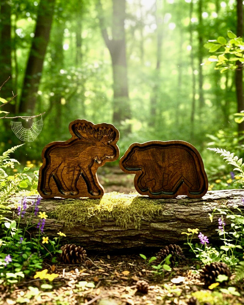 Two carved wooden animal figures, shaped like a moose and a bear, displayed side by side on a mossy log in a sunlit forest clearing.