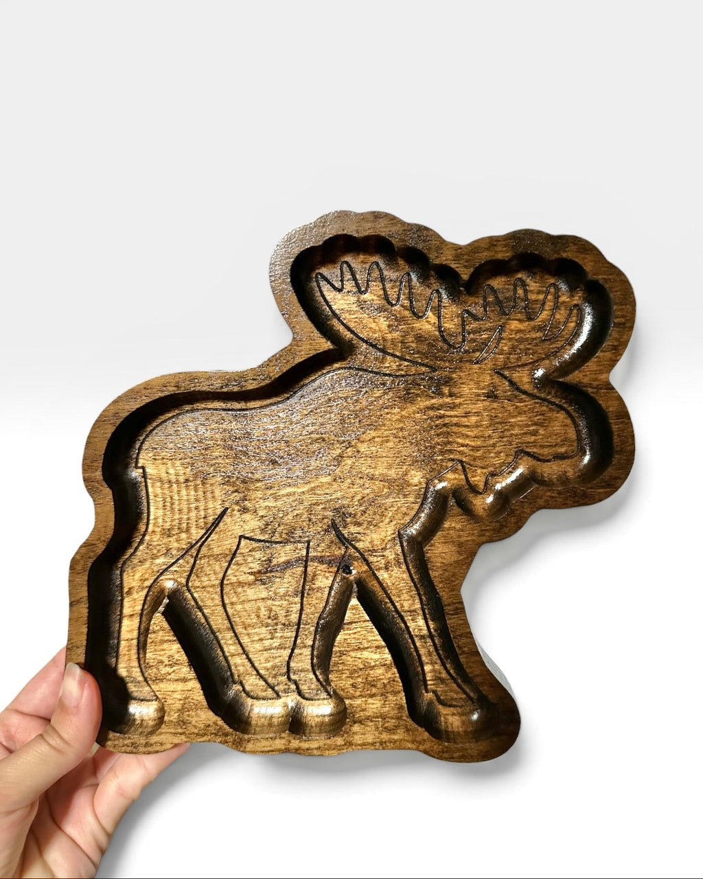 A hand holding a carved wooden moose-shaped tray or bowl with a rich brown wood grain, set against a plain light gray background.