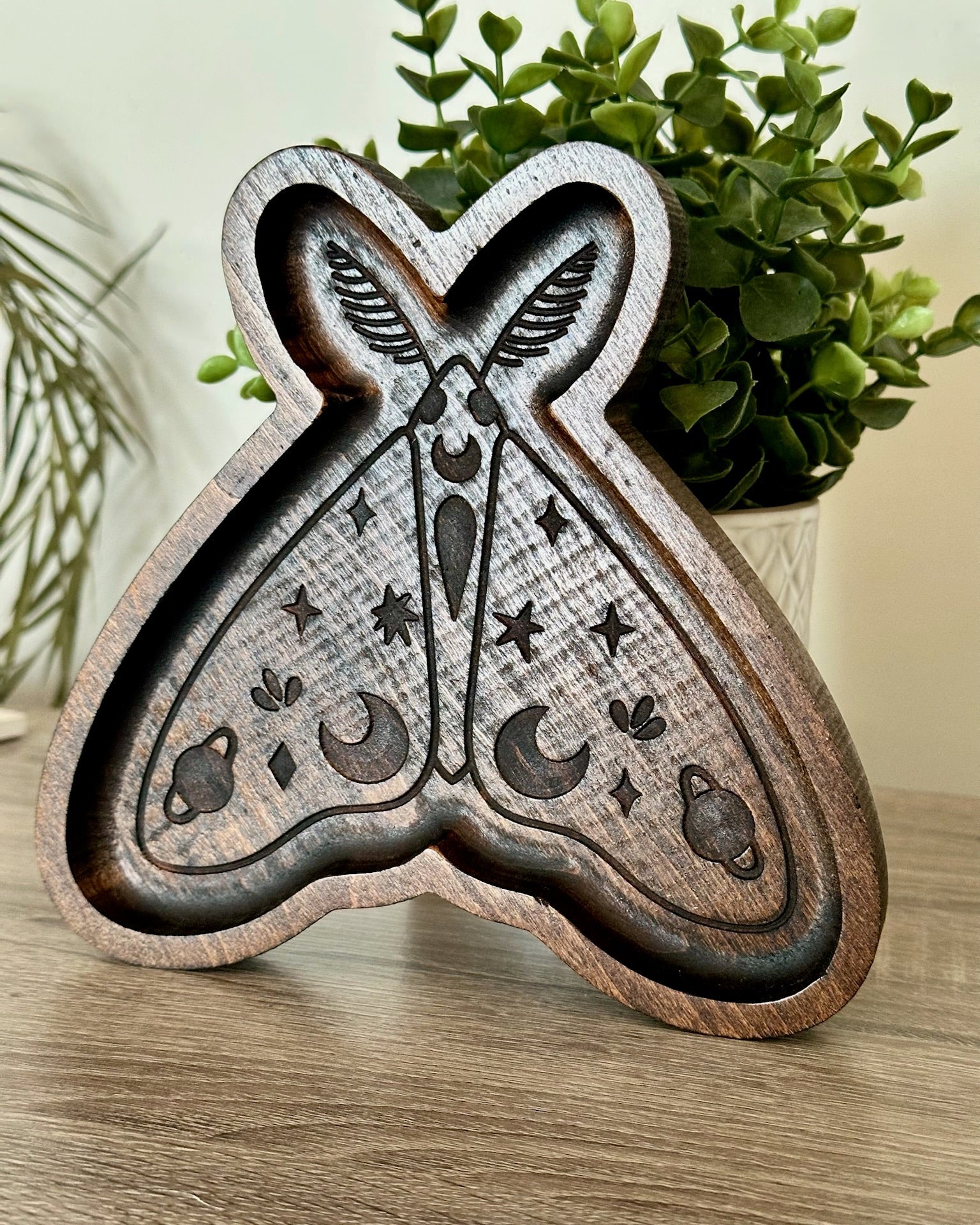 A carved wooden moth-shaped tray with celestial designs, displayed on a wooden tabletop with a green plant in the background.