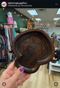 A hand with purple nail polish holds a dark brown carved wooden dish shaped like a mushroom in a retail store setting.