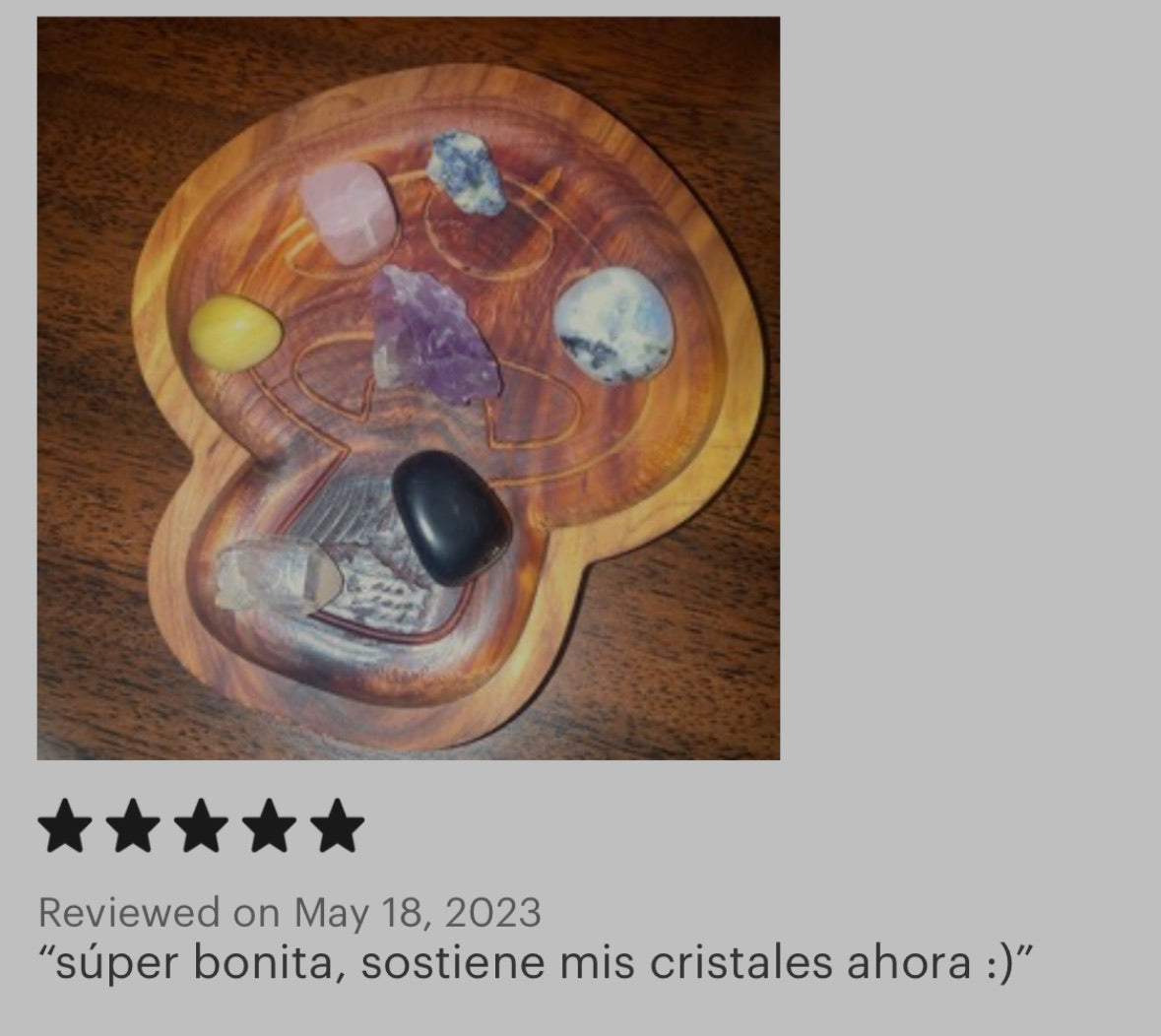 A heart-shaped mushroom tray holding several polished crystals and stones, shown on a dark wood surface. A review text appears below with a five-star rating and a comment in Spanish.