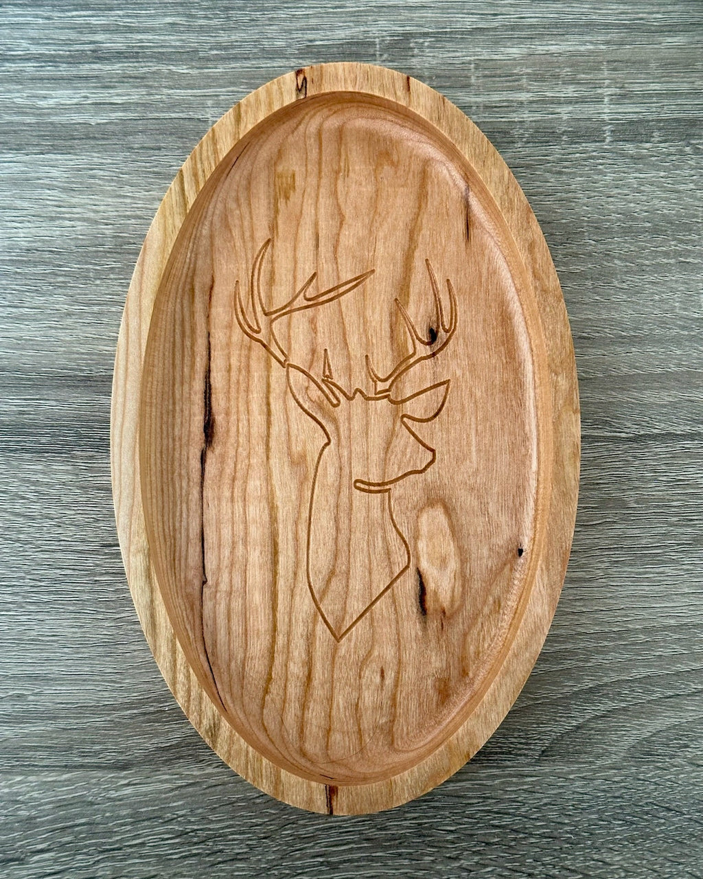 An oval wooden tray with a carved deer head design, displayed on a gray wood-textured background.